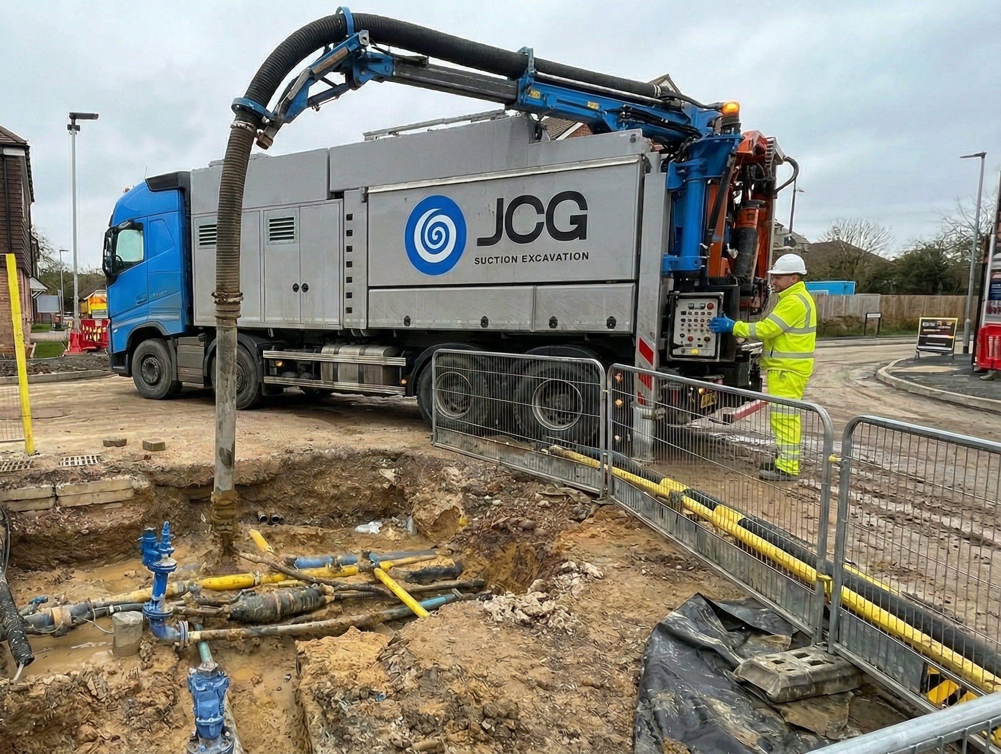 Construction worker operating a suction excavation truck at a construction site with exposed underground pipes and hoses.