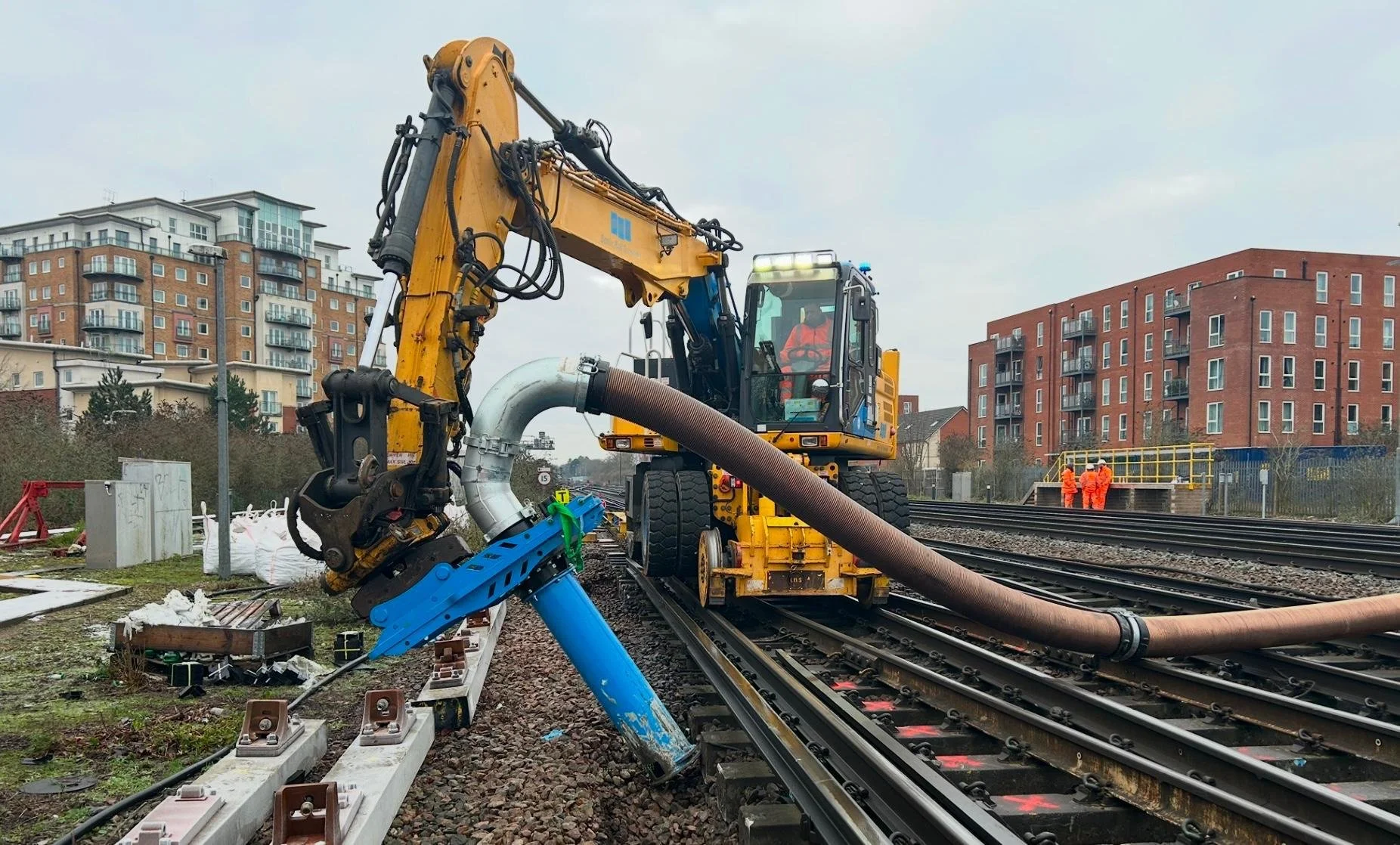 Construction workers and machinery working on railway track installation or maintenance, with buildings in the background.