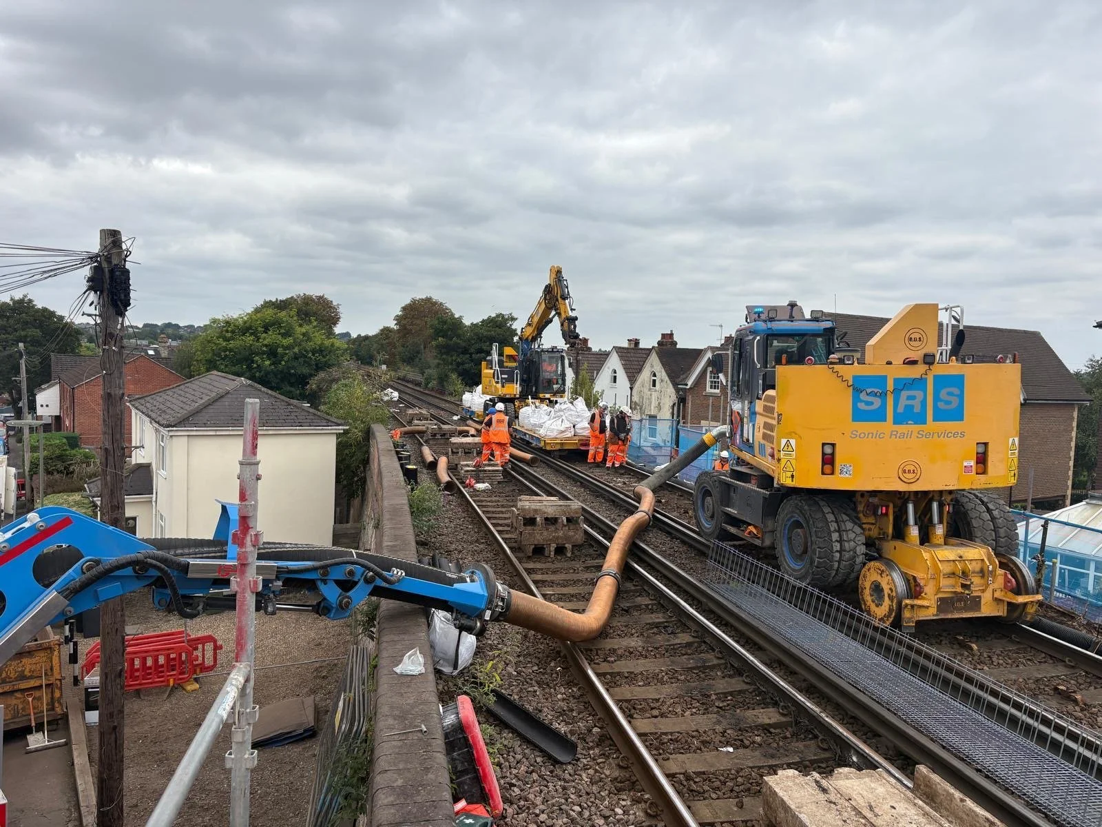 Construction workers operate machinery on a railway track near residential houses under cloudy skies.