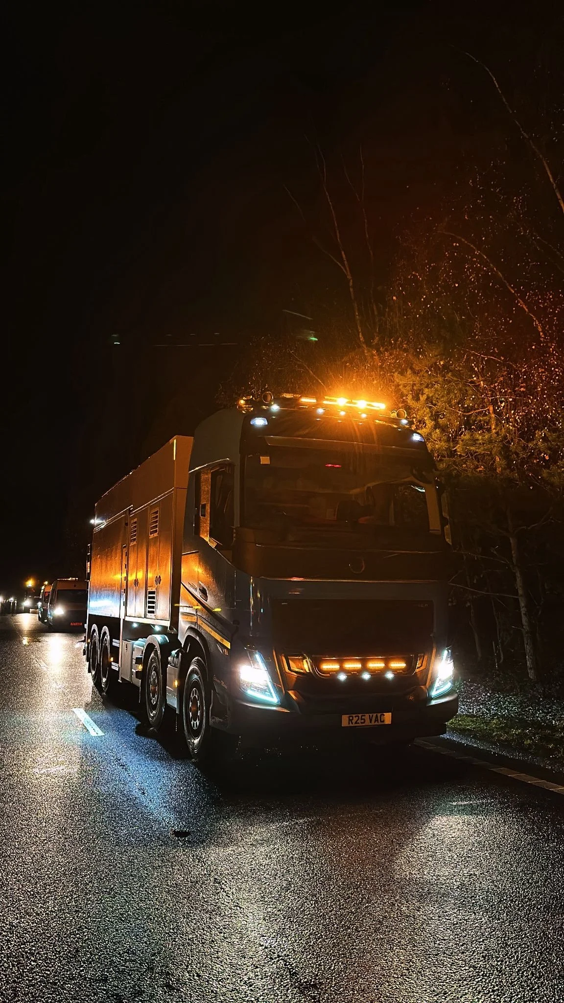 A large black utility truck with orange warning lights on the roof parked on a wet road at night, with trees and other vehicles in the background.