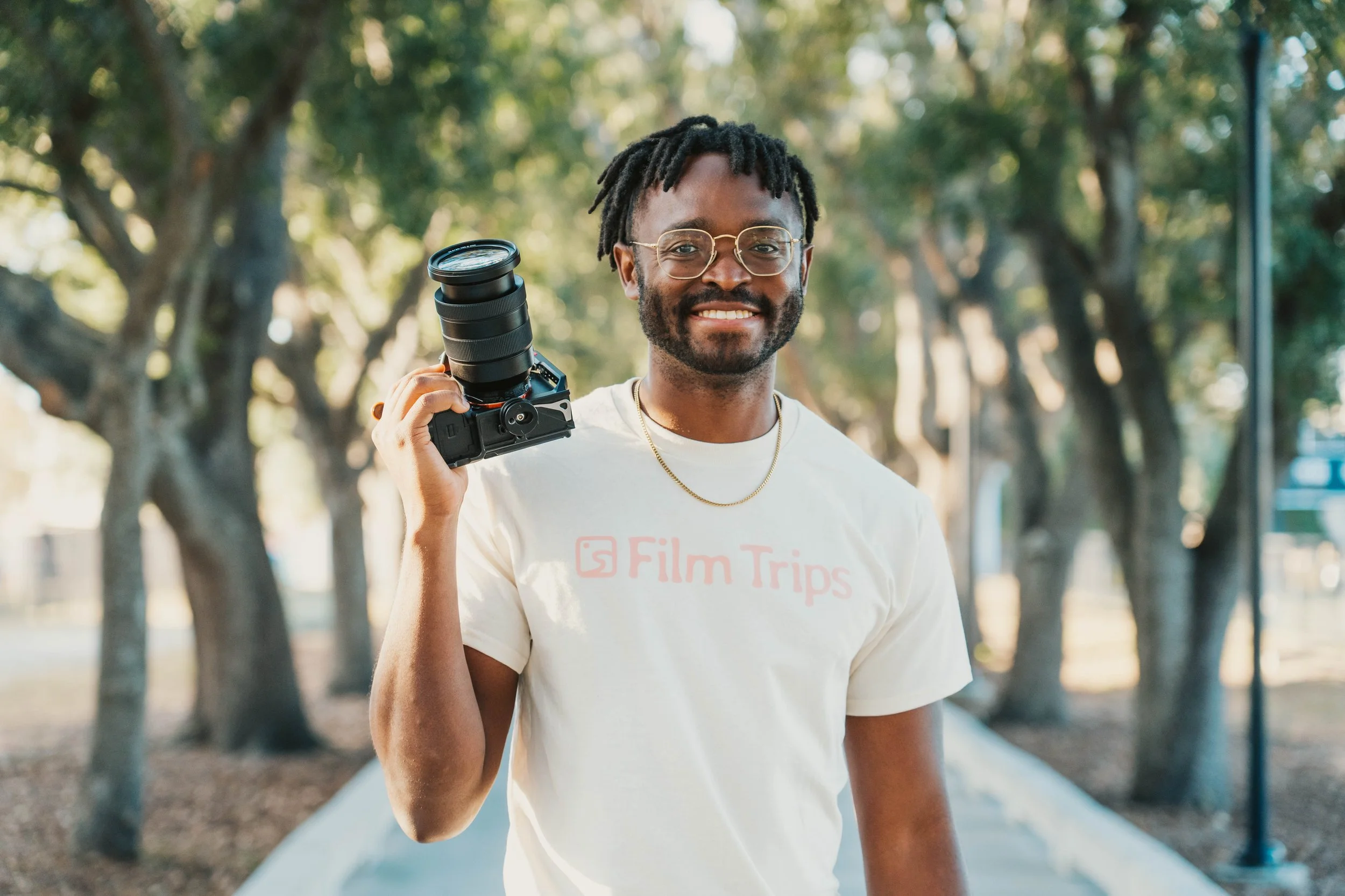 A young man with glasses and a beard, smiling, holding a camera with a zoom lens over his shoulder, outdoors in a park with trees and sunlight.