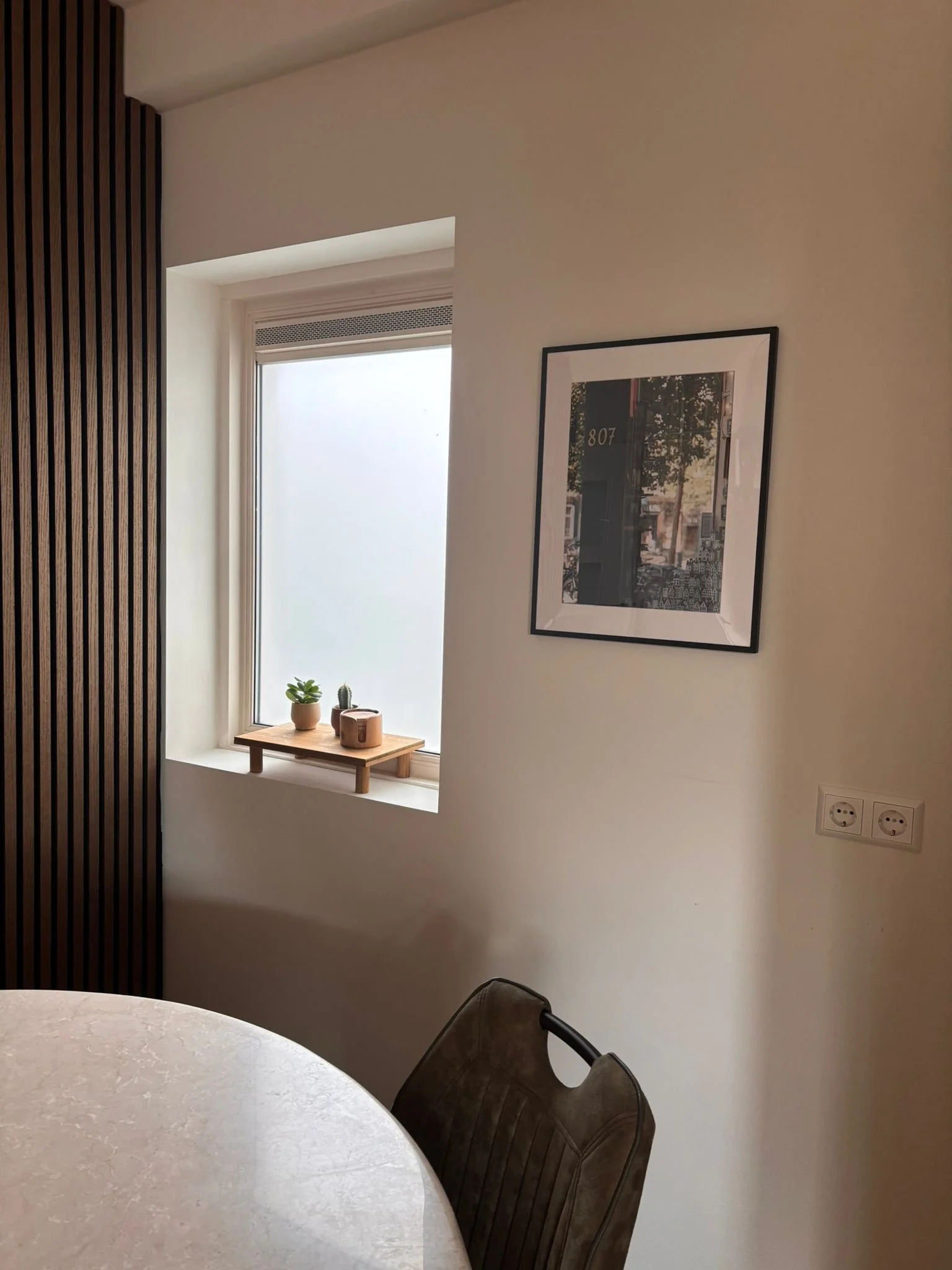 Een lichte kamer met een raam met jadegroene planten en decoratie op de vensterbank, een zwart-wit ingelijst foto aan de muur, en een ronde tafel met een bruine stoel.