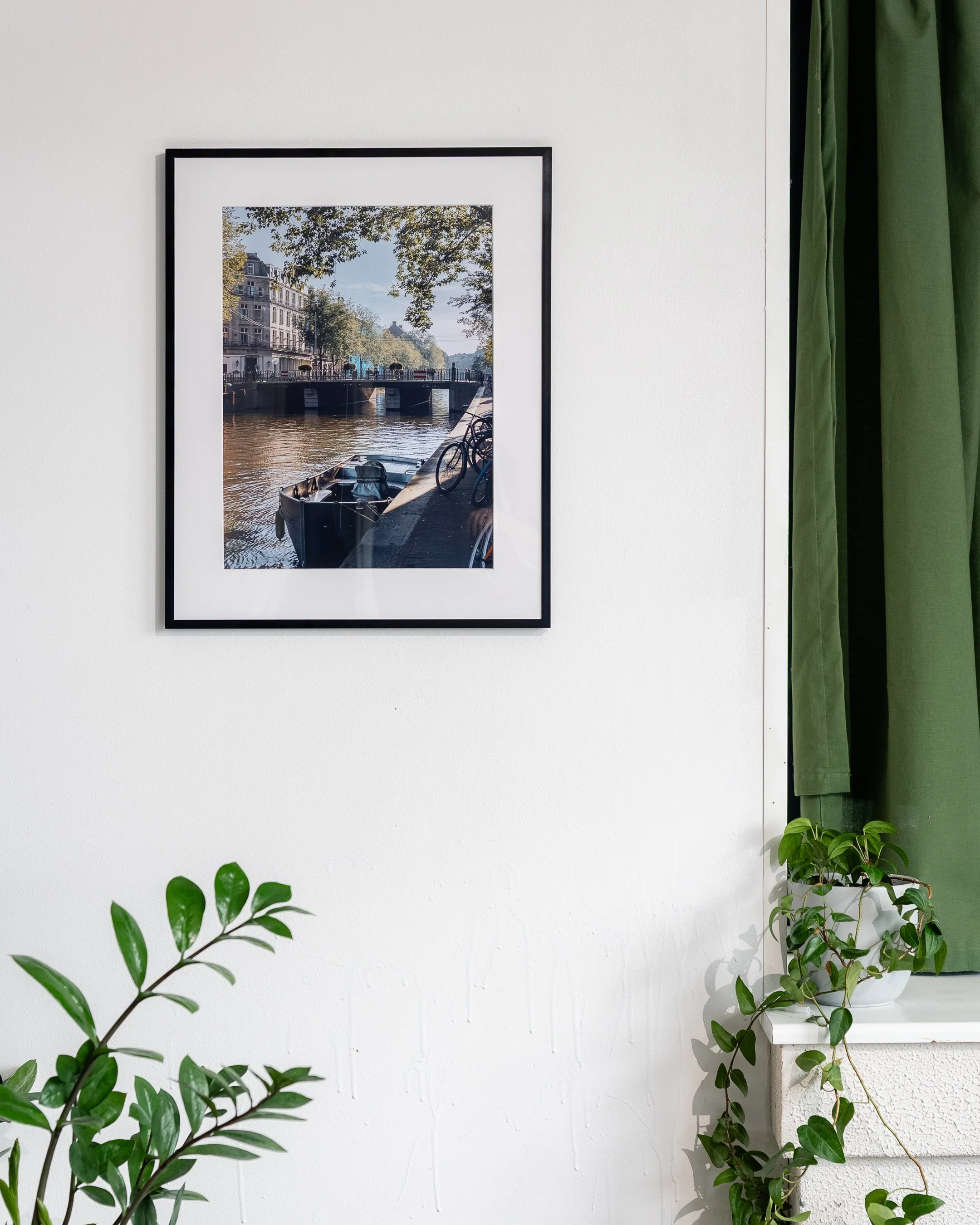 Ingelijste foto van een Amsterdamse gracht, witte muur, groene gordijnen, kamerplanten op de vensterbank.