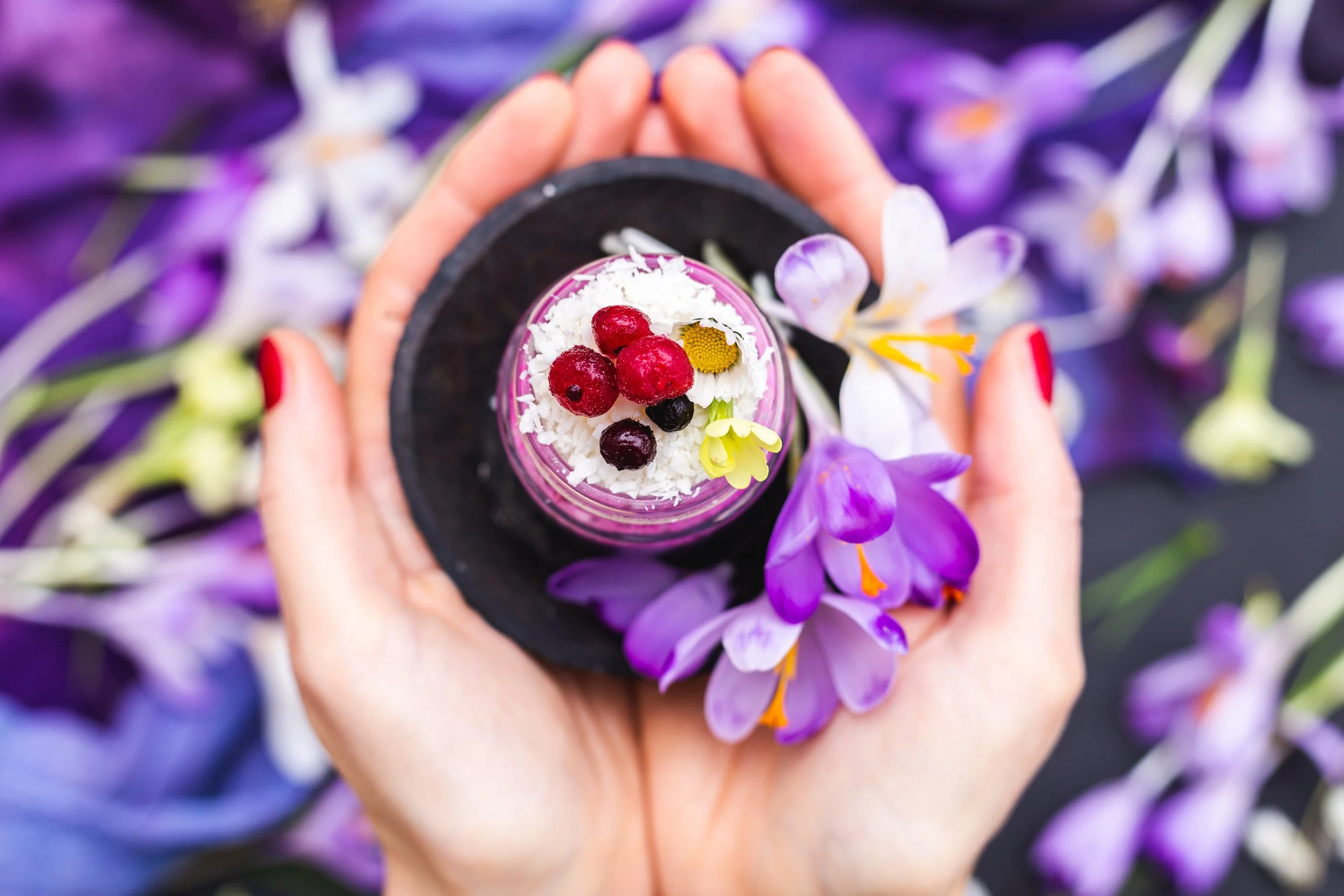 Une main tient une petite tasse contenant un dessert blanc avec des fruits rouges et noirs, des fleurs de camomille, entourée de fleurs de crocus violets et blanches, avec un fond flou de fleurs violettes.