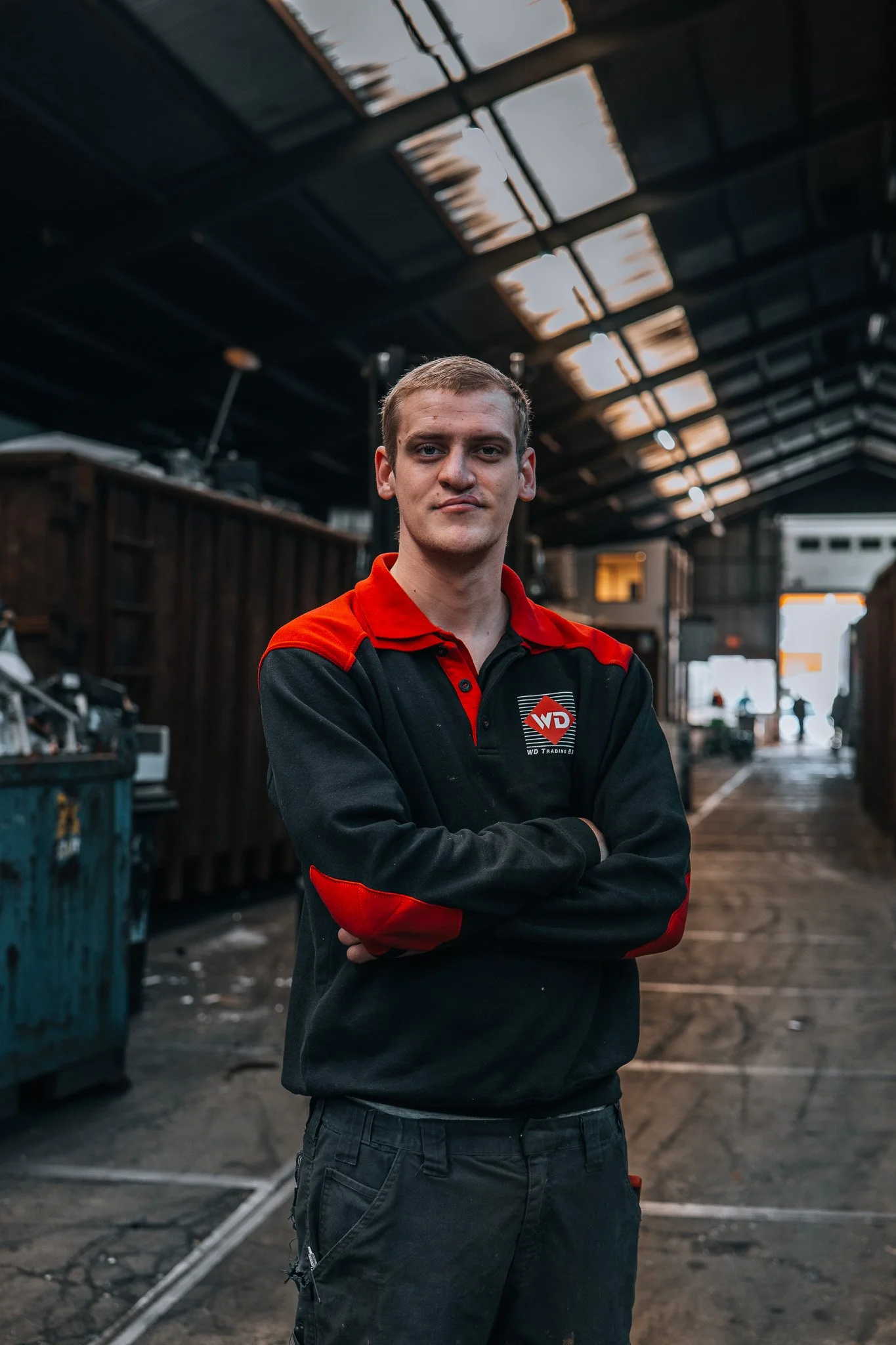 Een jonge man, loodsmedewerker, in een zwarte en rode werkoutfit met het logo 'WD Trading' staat met gekruiste armen in een loods, werkplaats of magazijn in Krimpen aan den IJssel