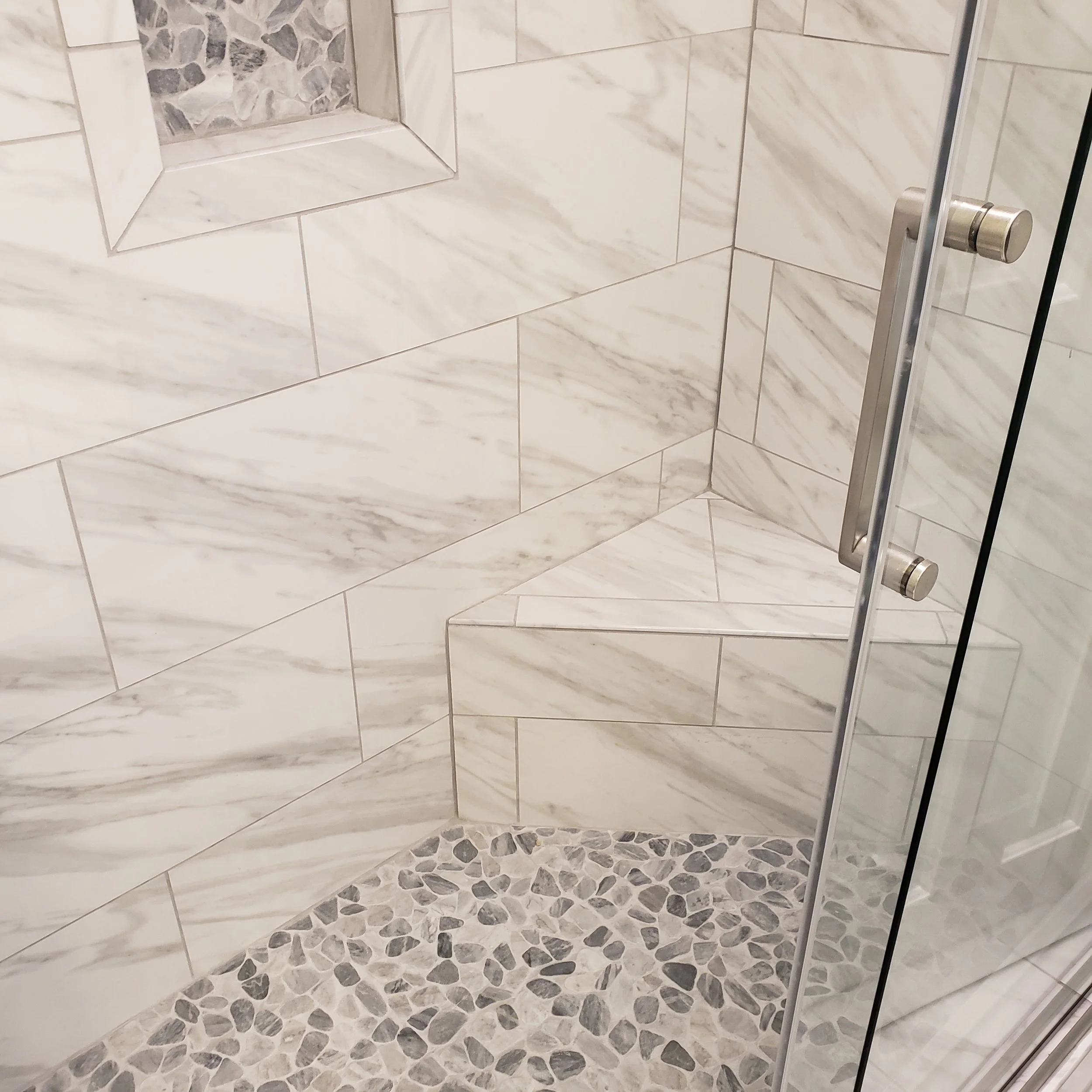 Close-up view of a marble tiled shower with a glass door, featuring a built-in niche with pebble flooring inside the shower.
