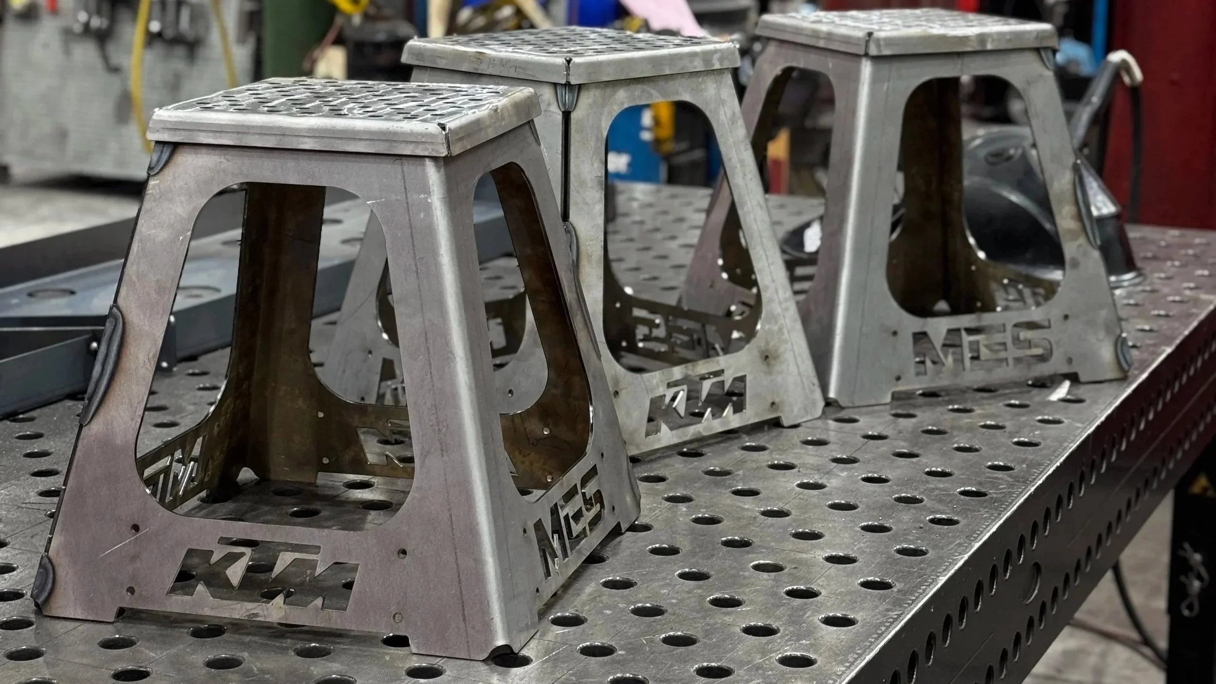 Three metal KTM bicycle kickstands placed on a perforated metal work surface in a workshop setting.