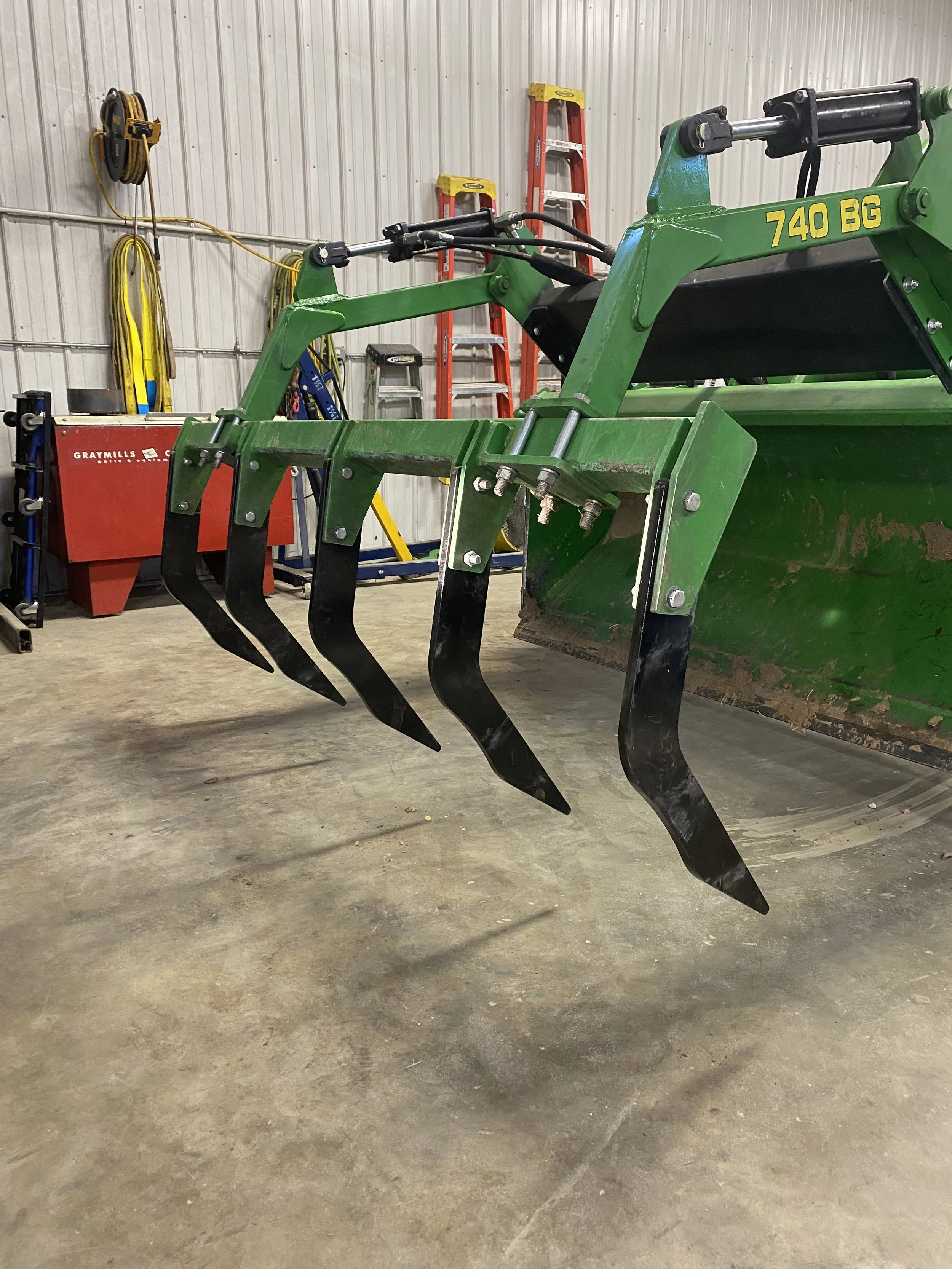John Deere front loader tractor with black tines in a garage or workshop with ladder and equipment in the background.