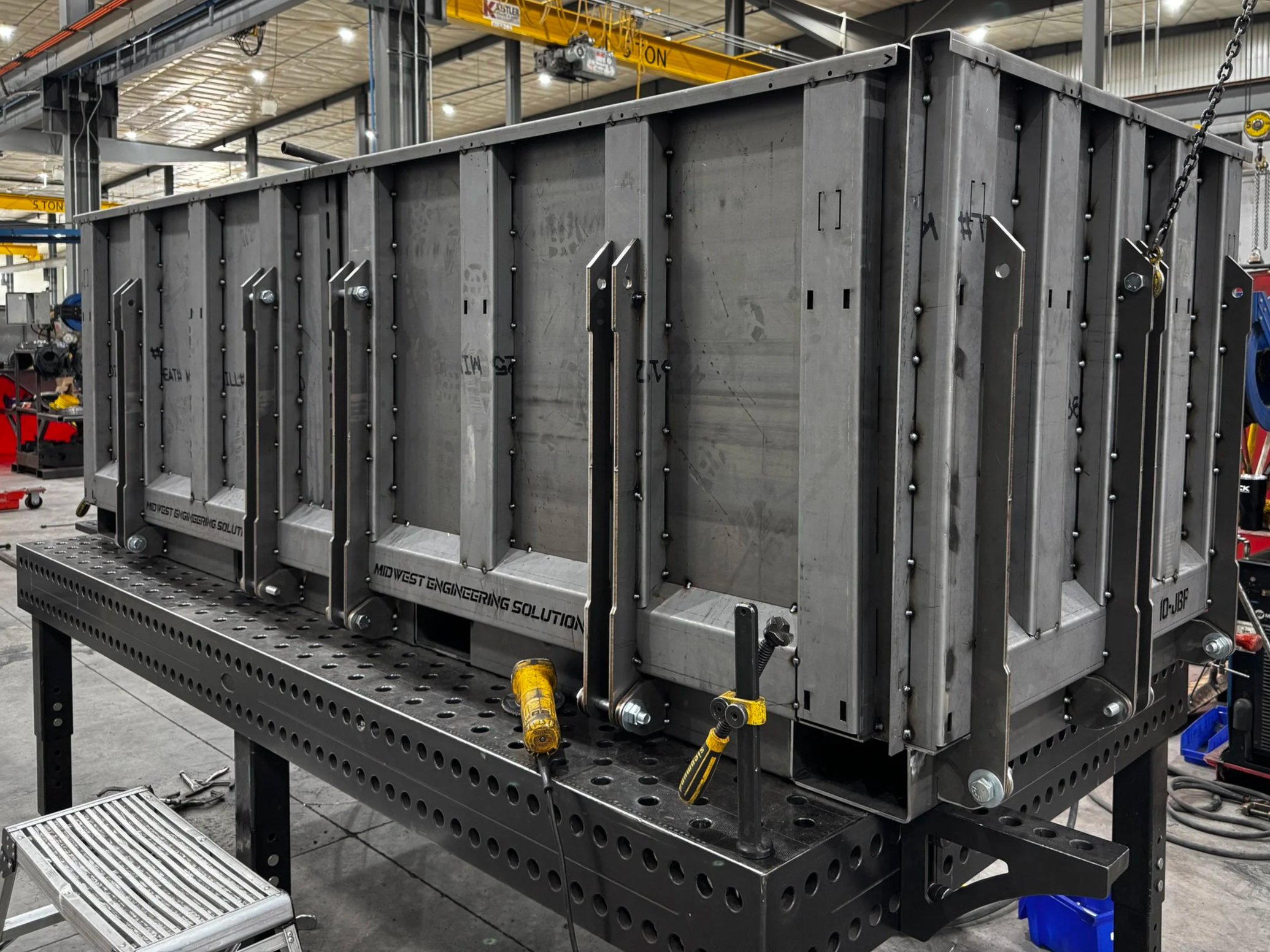 Large metal industrial structure on a worktable in a factory or workshop setting. The structure has multiple vertical panels secured with bolts and various tools and parts around it.