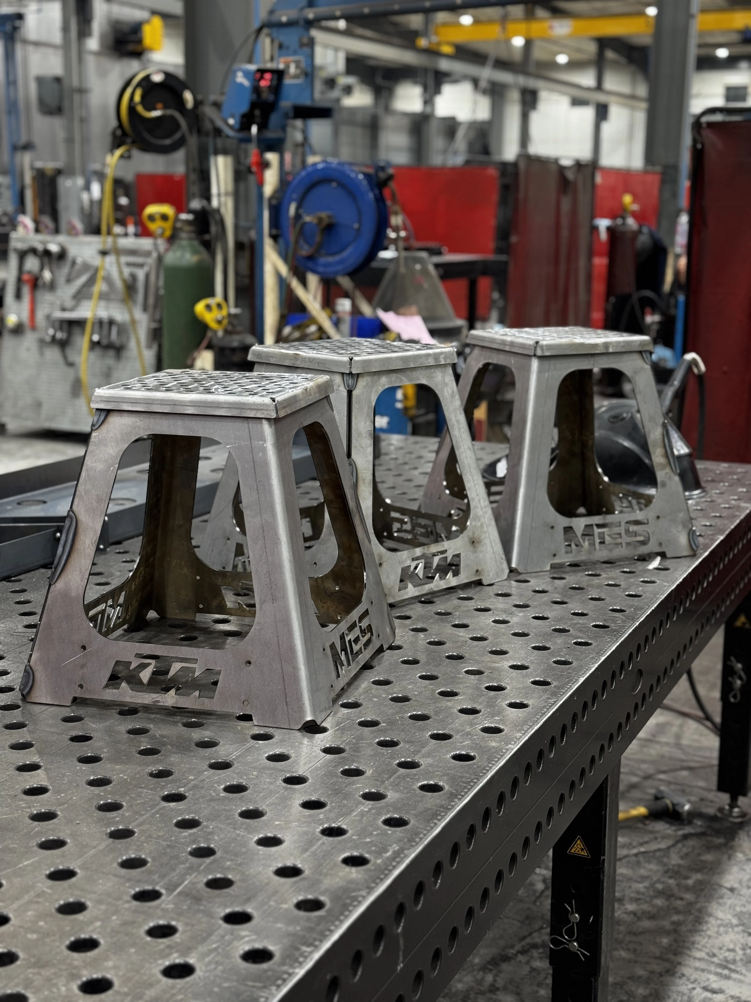 Three metal dirt bike stands with the KTM logo on the side, positioned on a perforated metal workbench in a workshop.