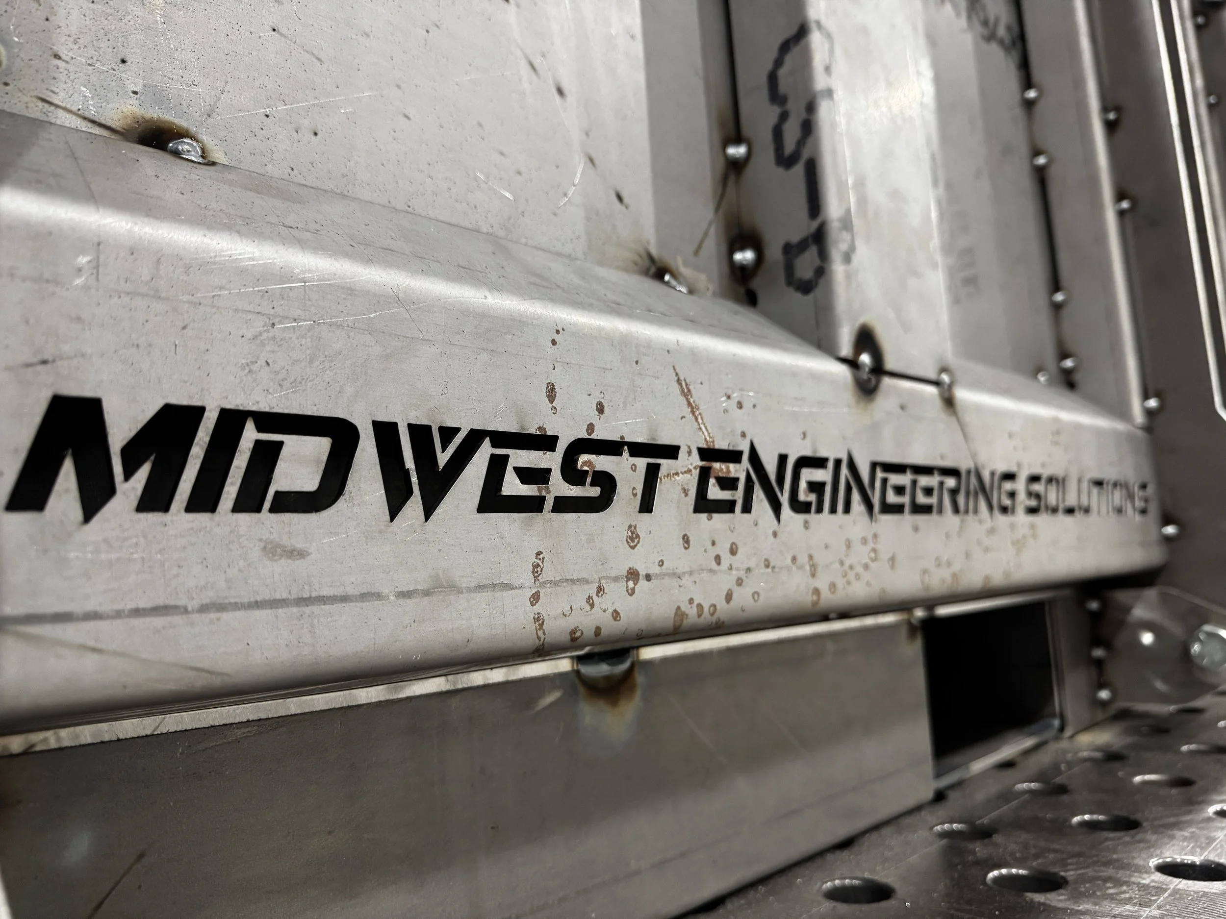 Close-up of a metal machine part with black lettering, some rust, and scrapes, showing a portion of the word 'ENGINEERING' and parts of other words, with visible screws, rust spots, and a worn surface.