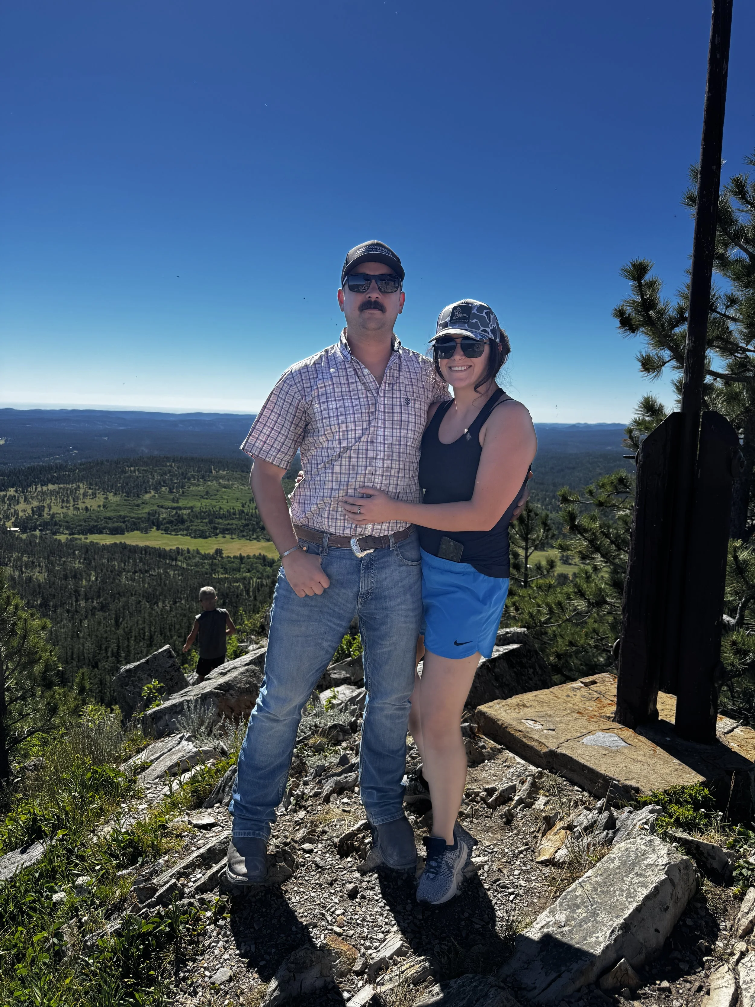 A man and a woman are standing outdoors on a rocky trail with a scenic view of a forested valley and mountains in the background. They are smiling, wearing sunglasses and casual outdoor clothing, and appear to be enjoying a hike on a sunny day.