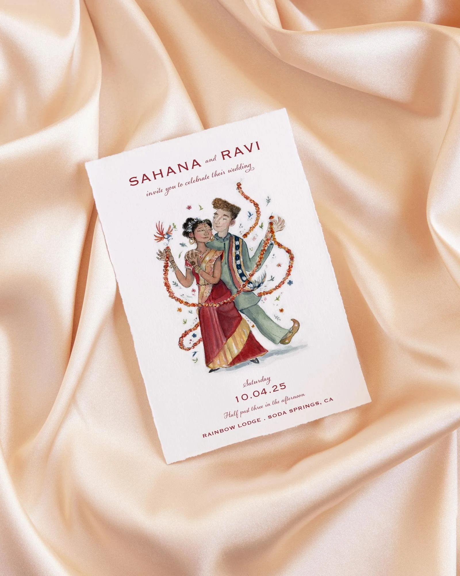 Wedding invitation with an illustration of a couple in traditional Indian attire, celebrating their wedding. The invitation is placed on light peach satin fabric.