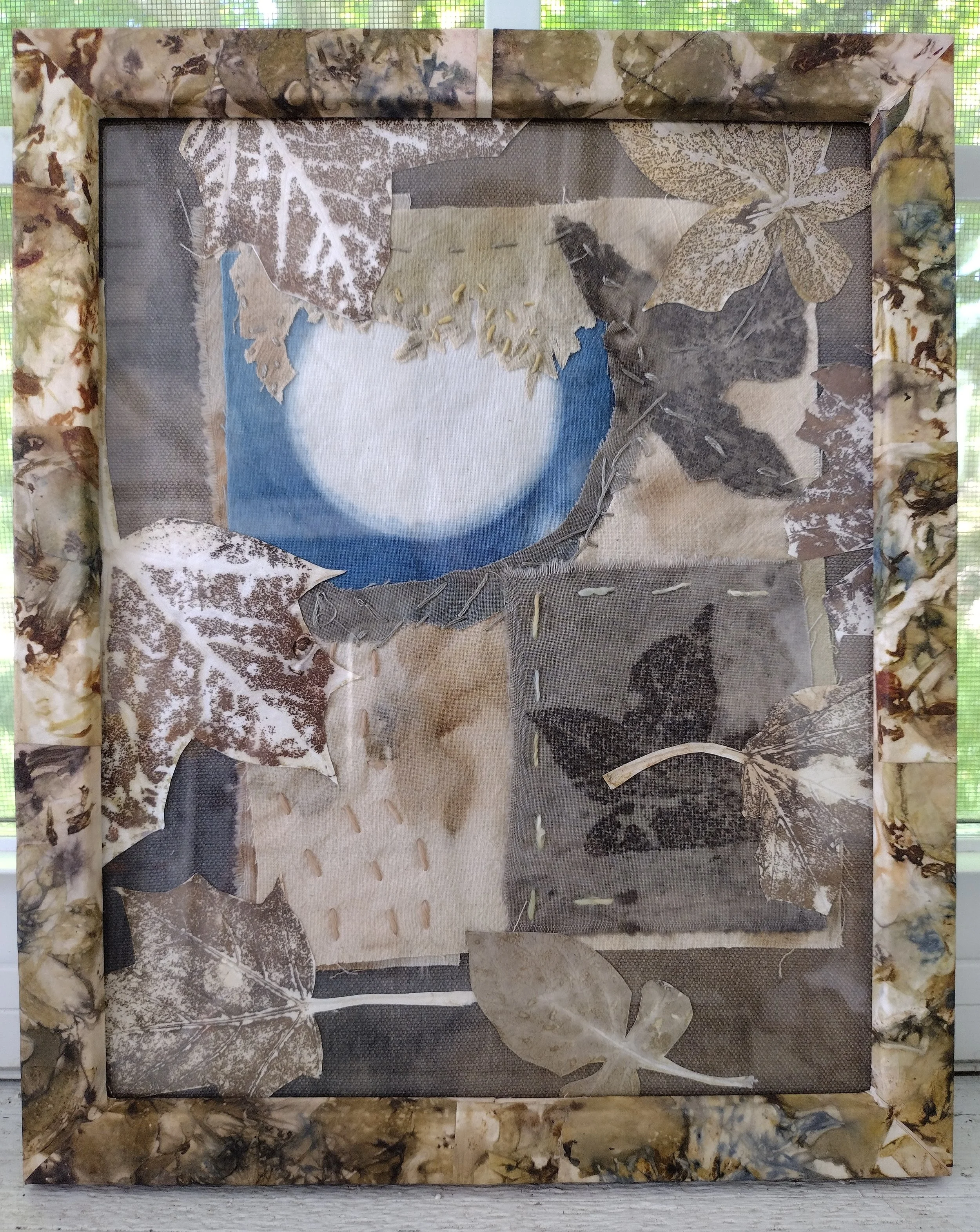 A shadow box display with a natural bark-like frame, containing various dried leaves and plant materials arranged on a background of different textured paper and fabric pieces, forming a botanical collage. By Amy Real