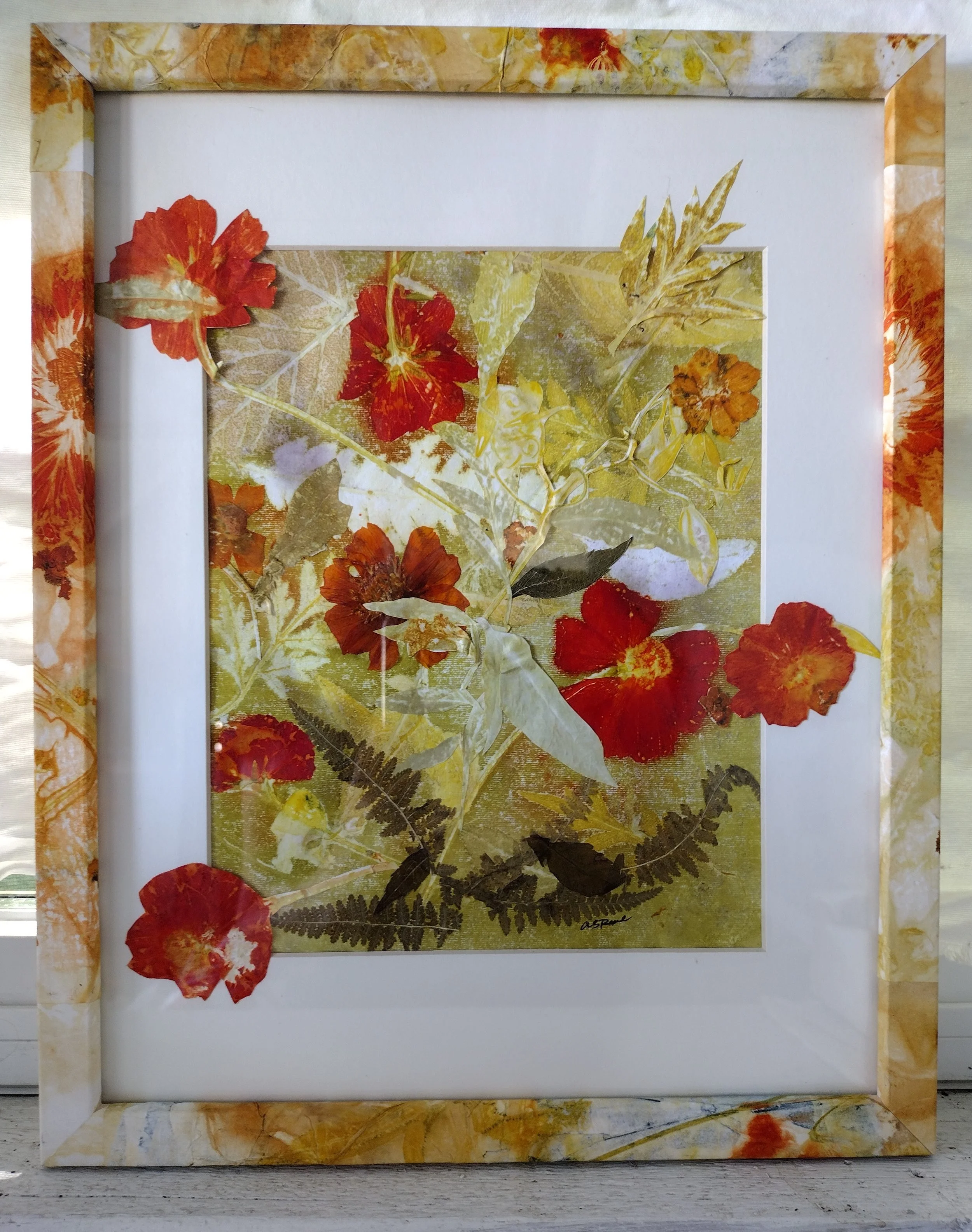 A decorative framed artwork featuring dried autumn leaves and flowers in shades of red, orange, yellow, and brown, arranged on a textured background. By Amy Real