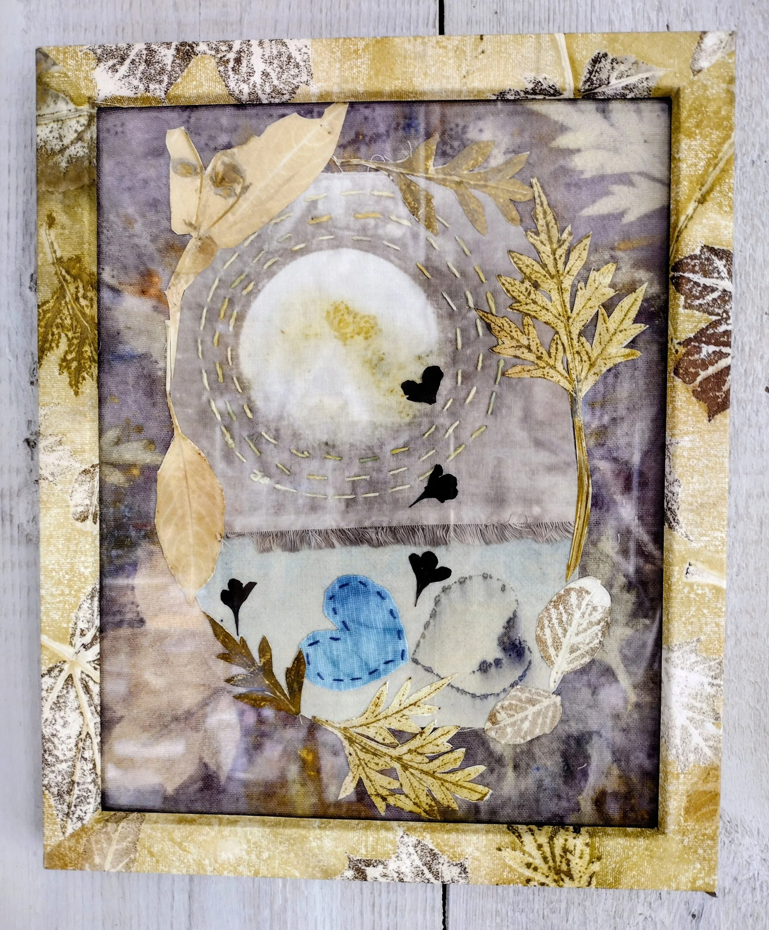A framed fabric art piece with a collage of leaves, hearts, and stitched designs, featuring a moon or sun element, on a neutral background with a gold and brown border. By Amy Real