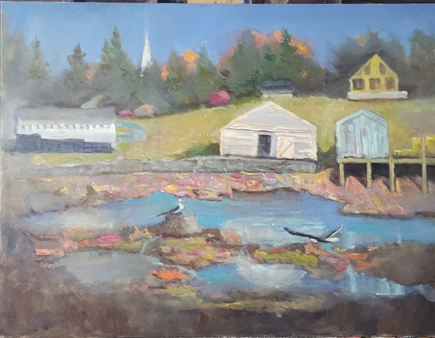 Oil painting of a seaside scene with a rocky shore and water in the foreground, several beach huts and buildings on a grassy hill, surrounded by trees with autumn foliage. by Patrick Monaghan