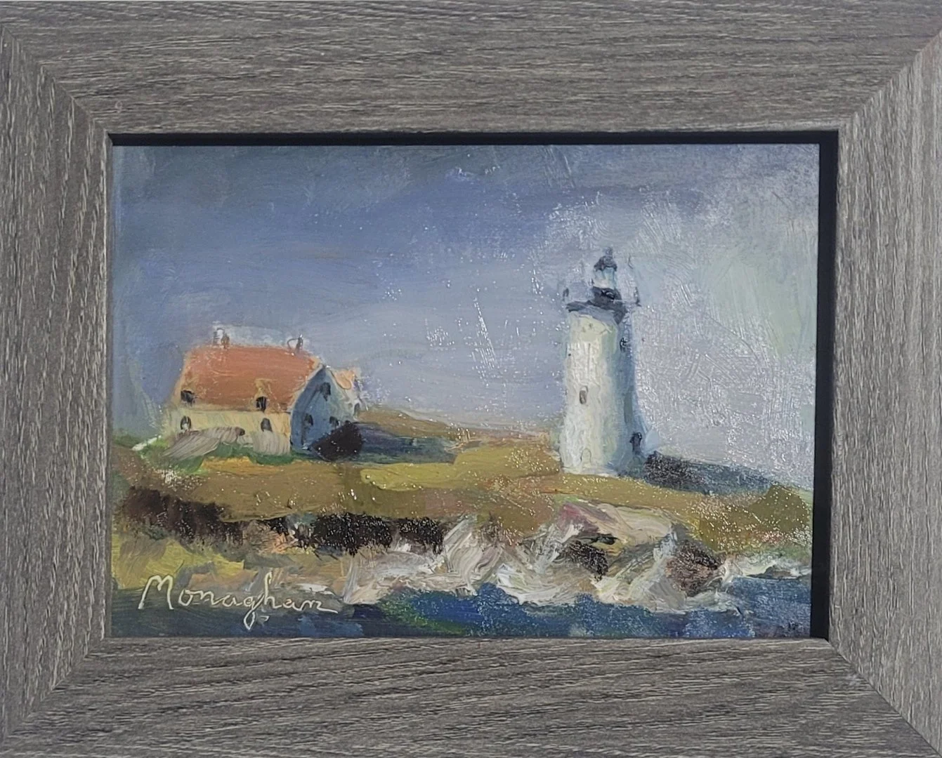 A framed landscape painting of a lighthouse and a house on a rocky coastline, signed 'Monaghan' in the bottom left corner.