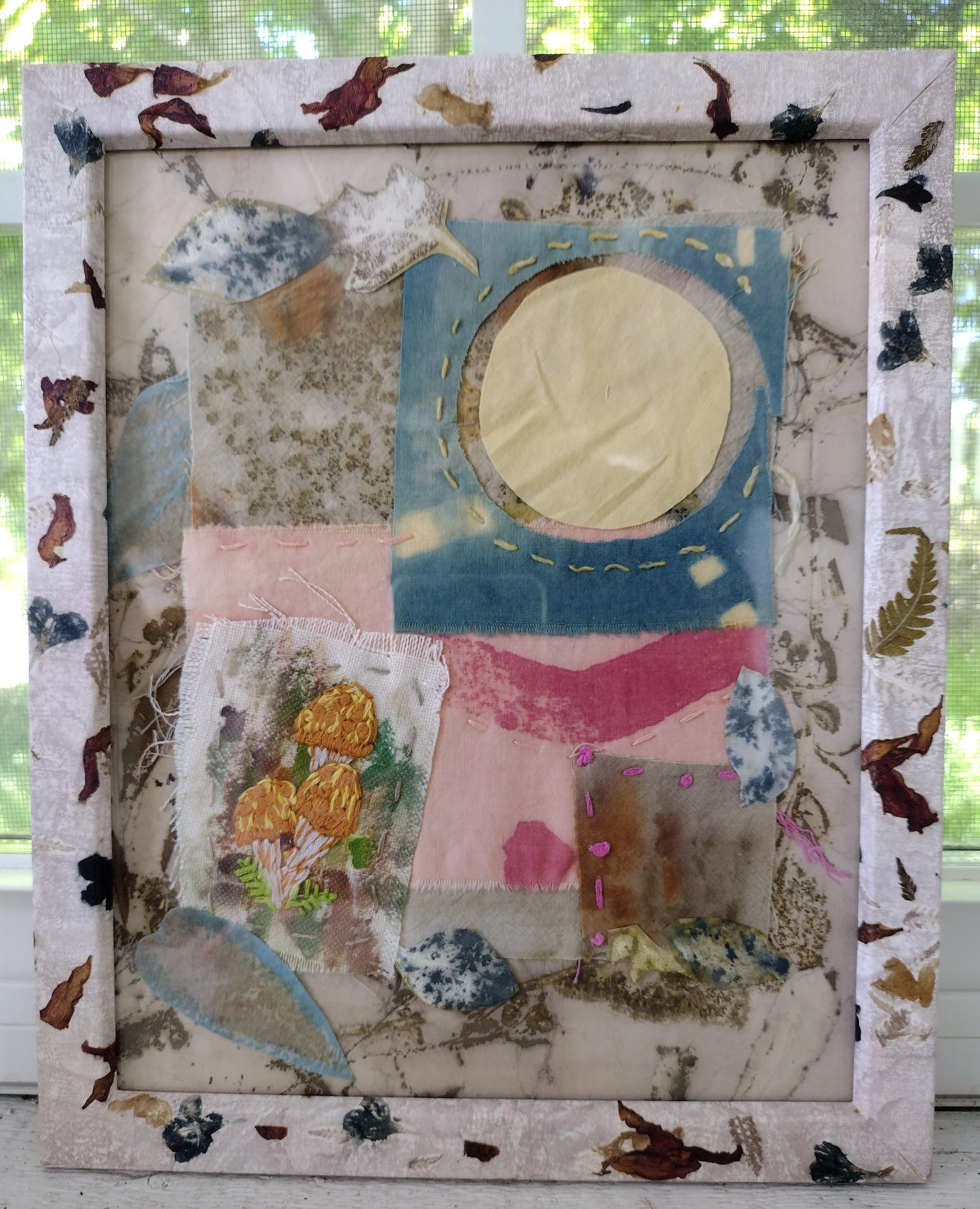 Mixed media artwork featuring leaves, fabric, and embroidery on paper, framed with pressed leaves and nature-inspired decorations. By Amy Real