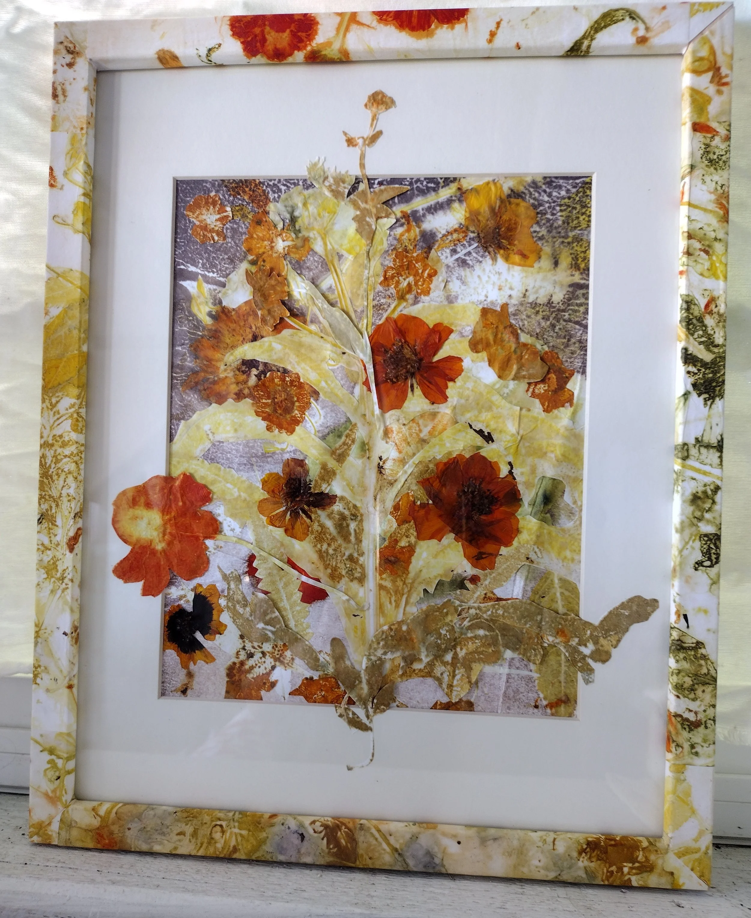Framed artwork featuring pressed flowers and leaves arranged in an abstract pattern, with a colorful floral border. By Amy Real