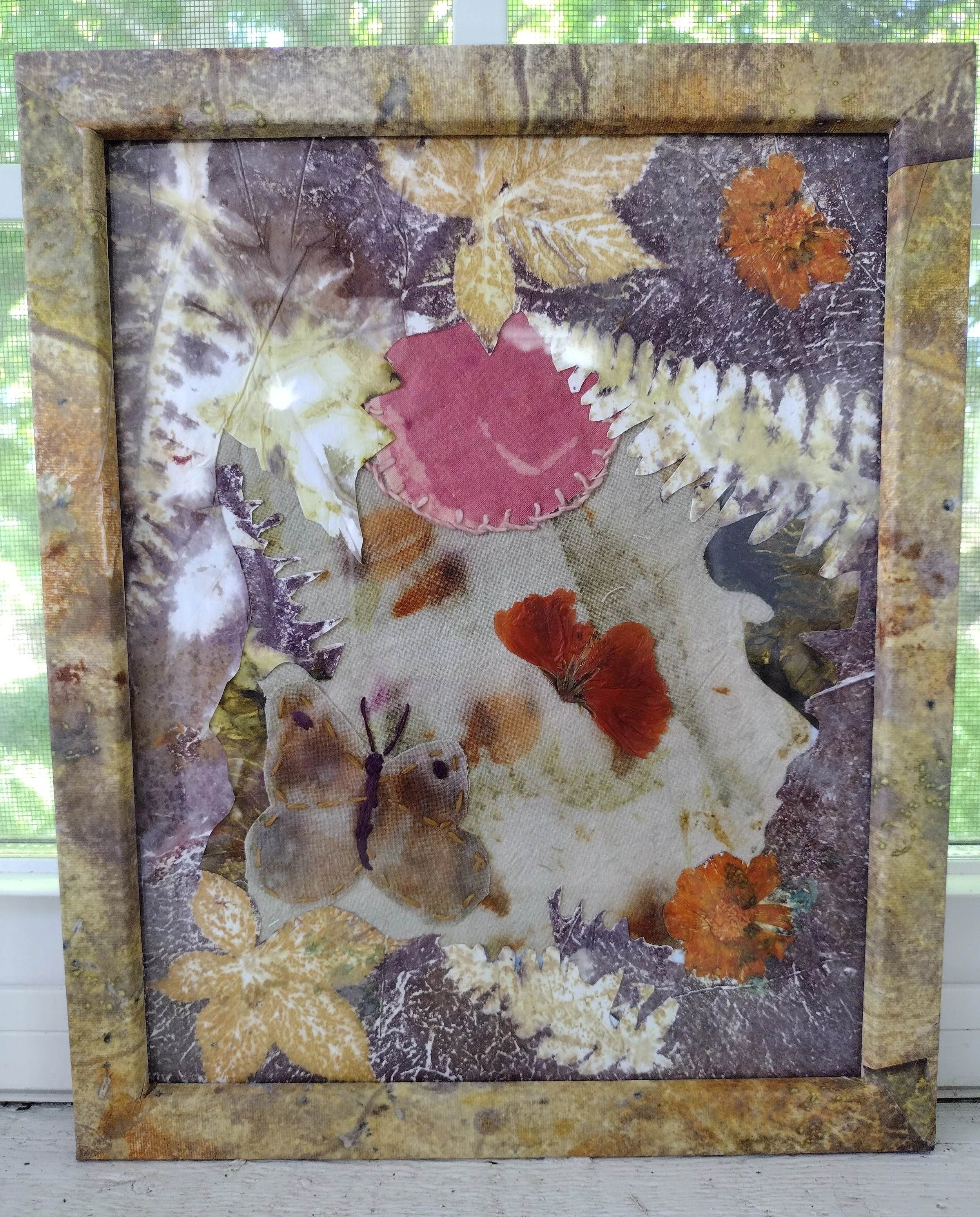 A framed piece of art depicting various dried leaves and flowers arranged on a textured background with a protective glass cover, sitting on a white wooden surface with a window and greenery in the background. By Amy Real