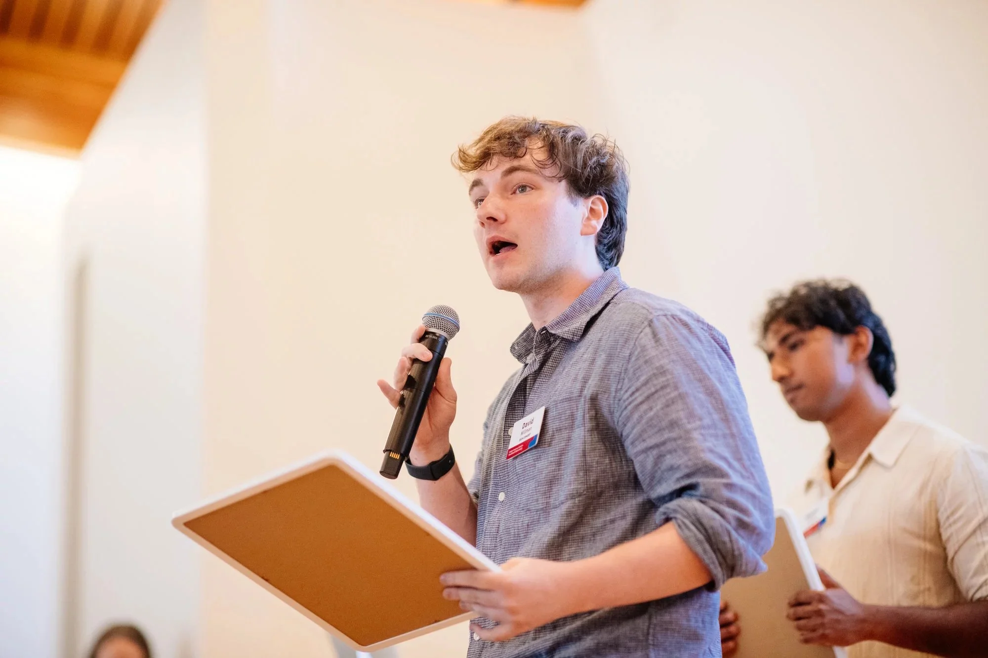 David Millman is speaking into a microphone while holding a clipboard during a presentation or speech. Another person is visible in the background holding a notebook.