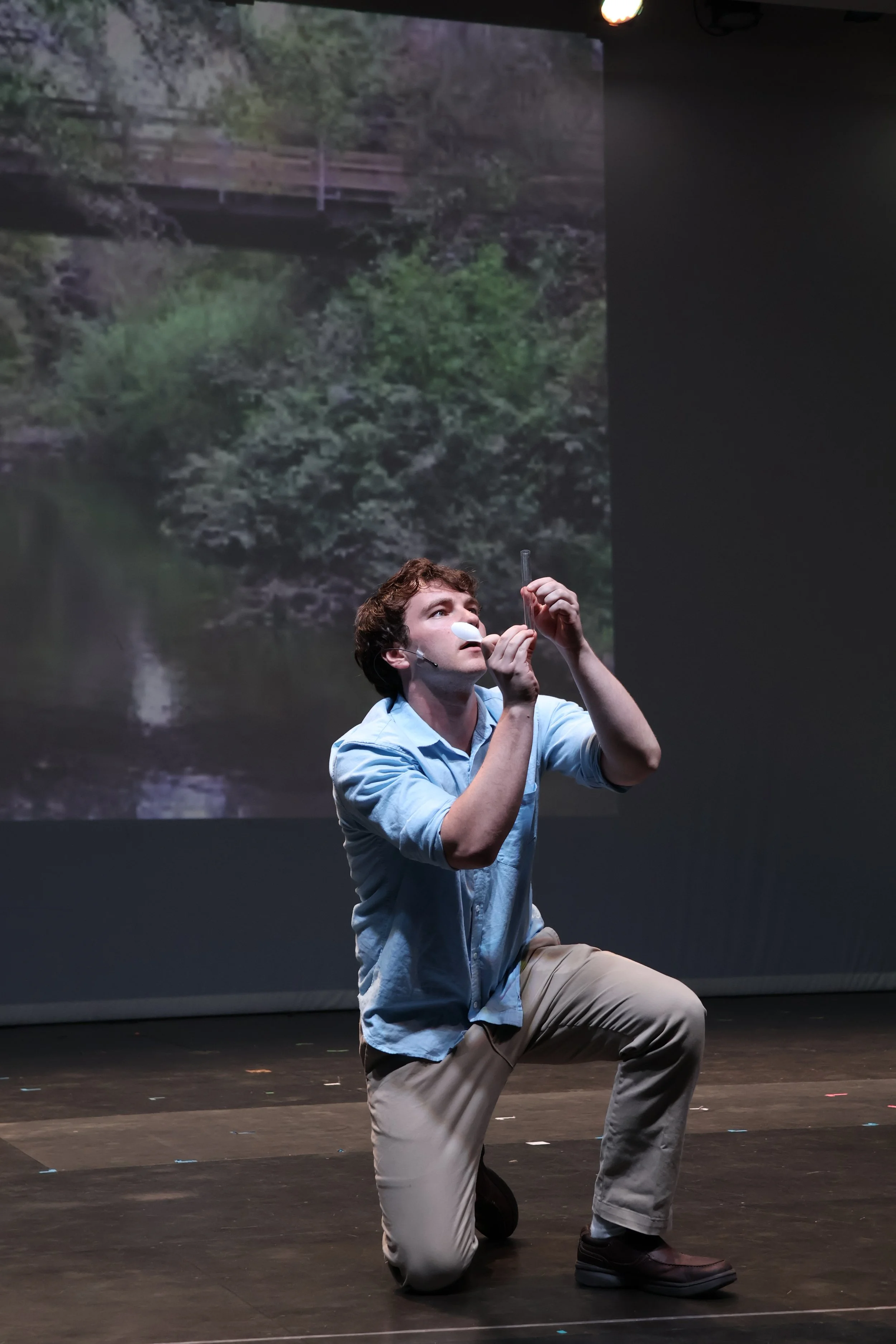 A young man kneeling on stage, holding a microphone and a test tube, with a nature scene backdrop of trees and a bridge.