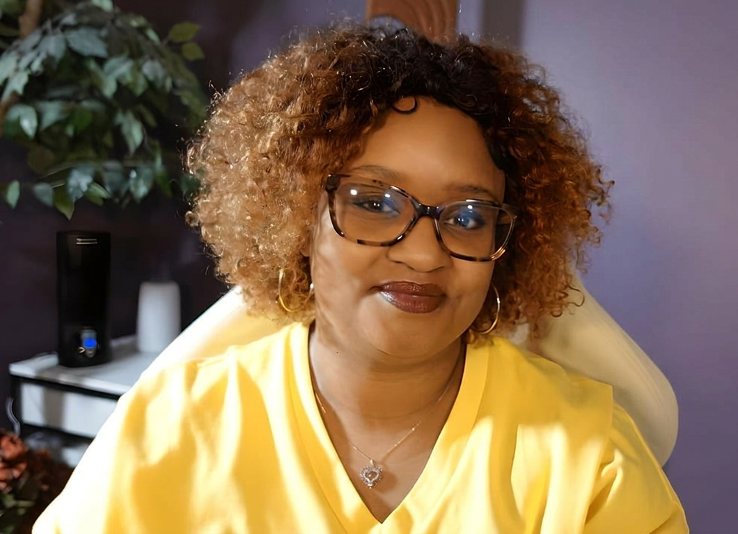 A smiling woman with curly, two-tone hair wearing glasses, gold hoop earrings, and a yellow shirt, sitting indoors in front of a plant and a side table with decor items.