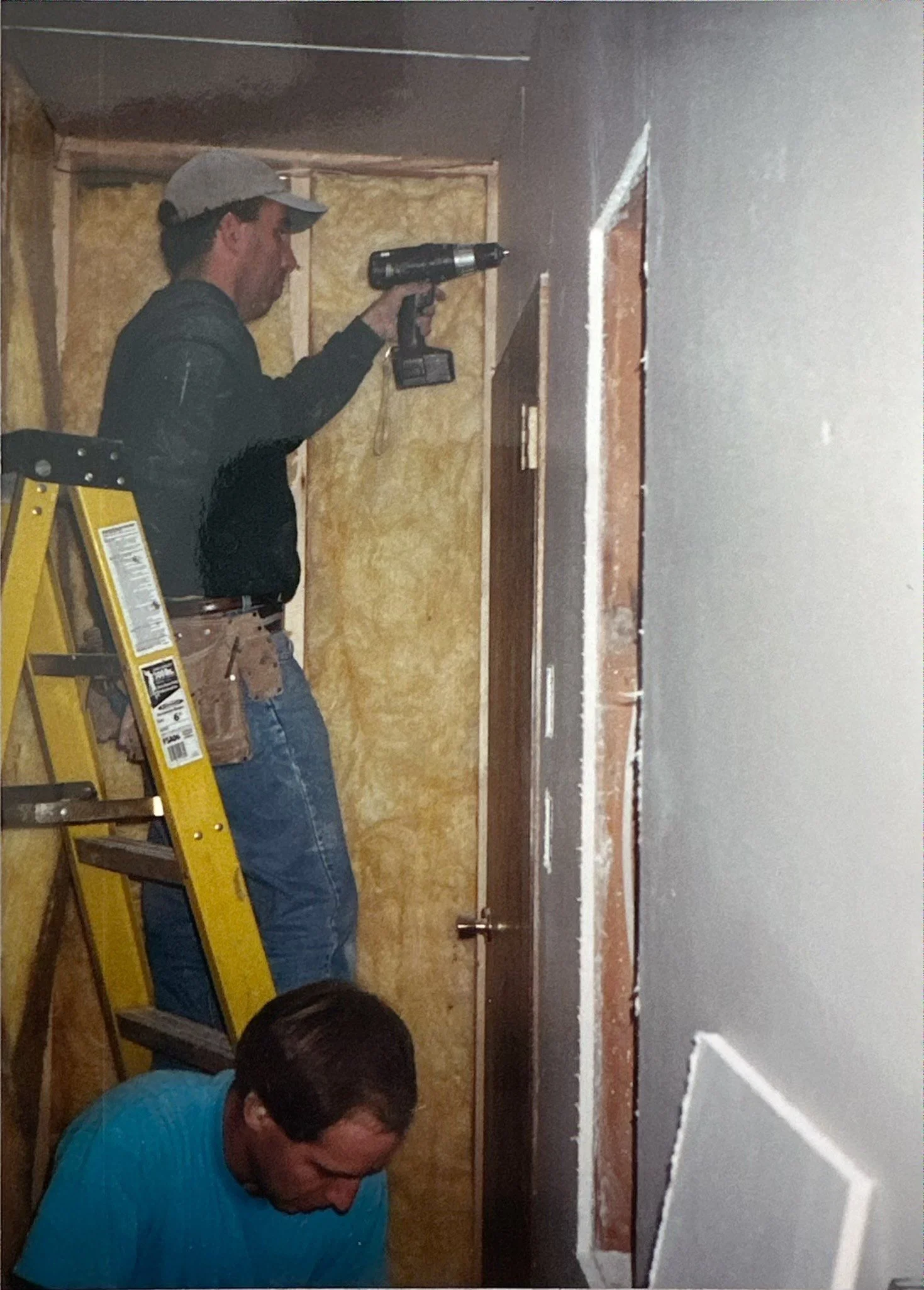 Matt Shaw, owner of Jay-Kay Construction working on a home on a ladder using a drill. The wall is partially unfinished with insulation visible.