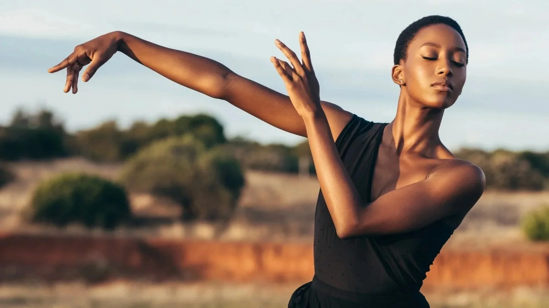 A woman with short hair dancing outdoors, with her eyes closed and arms extended, wearing a black sleeveless top.