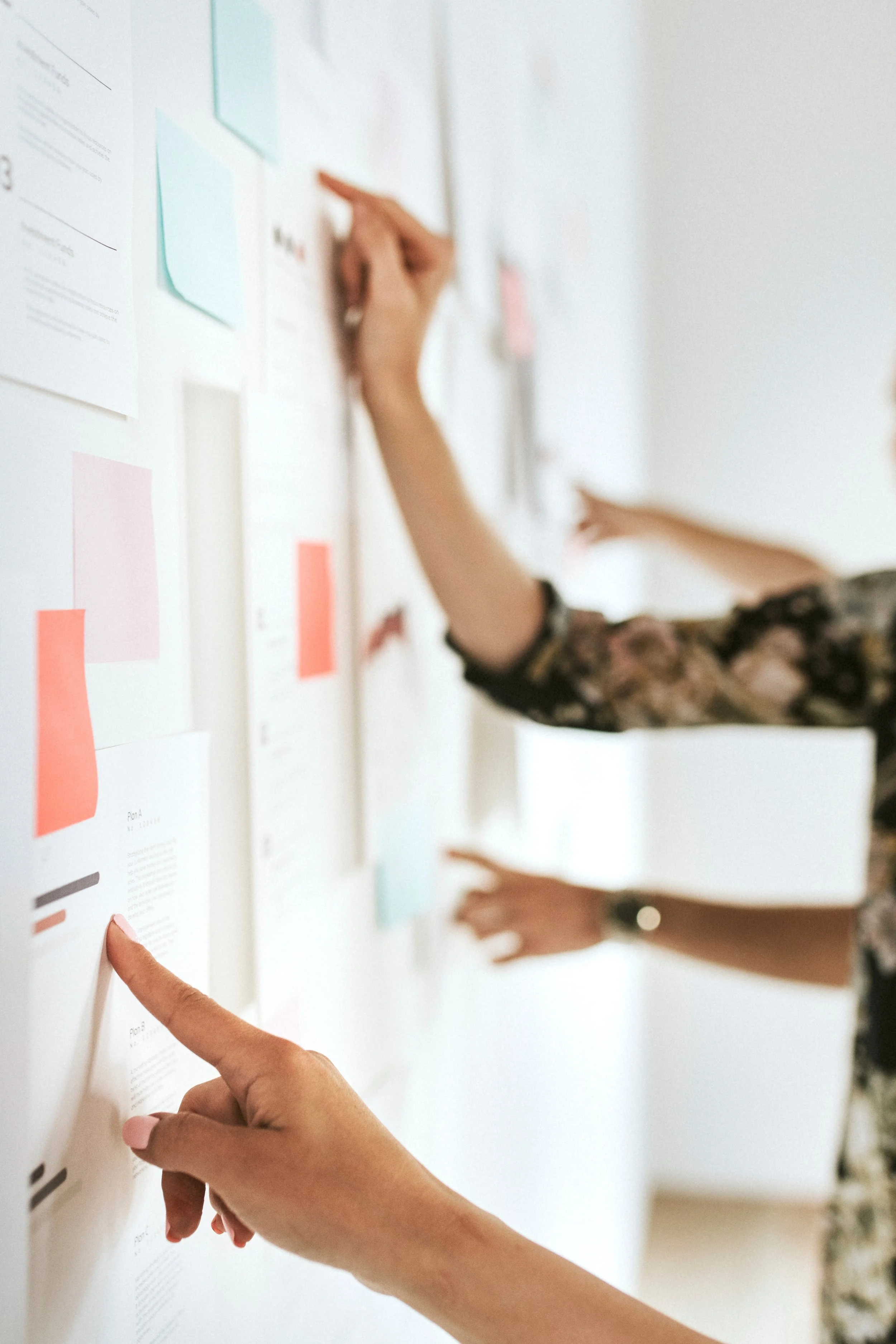 A person points at a document on a white wall with their index finger, while another person points at a different document, both engaged in a discussion or review.