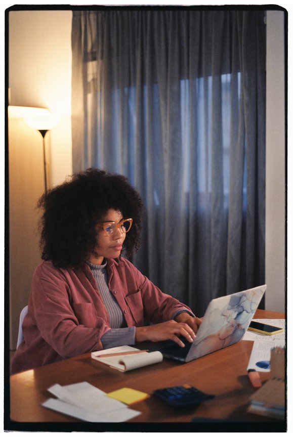 A woman with curly hair and glasses working on a laptop at a desk in a dimly lit room with curtains and a standing lamp.