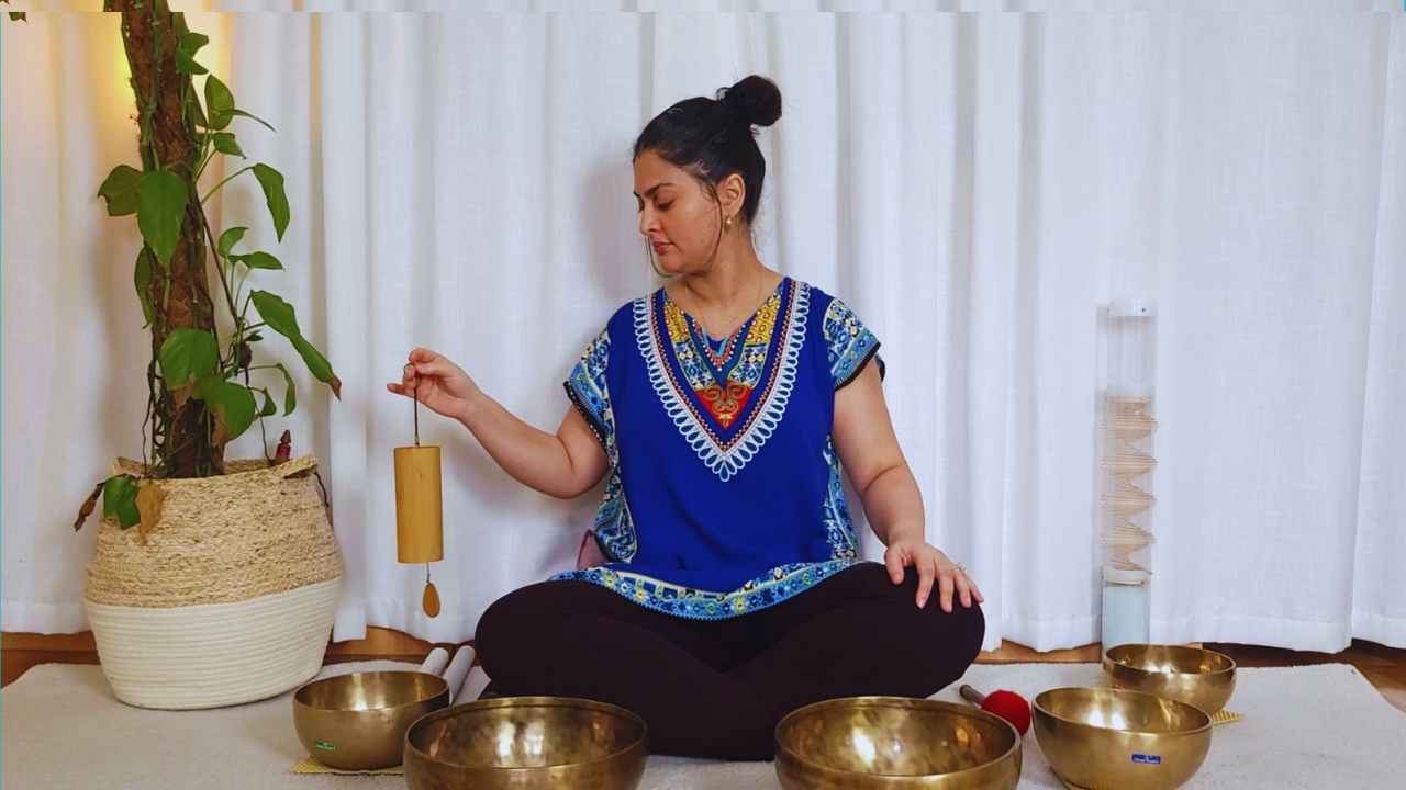 Sana sits cross-legged on the floor in a meditative pose, holding a chime in her right hand while surrounded by seven singing bowls. She is in a room with white curtains, a potted plant, and a decorative stacking crystal tower.