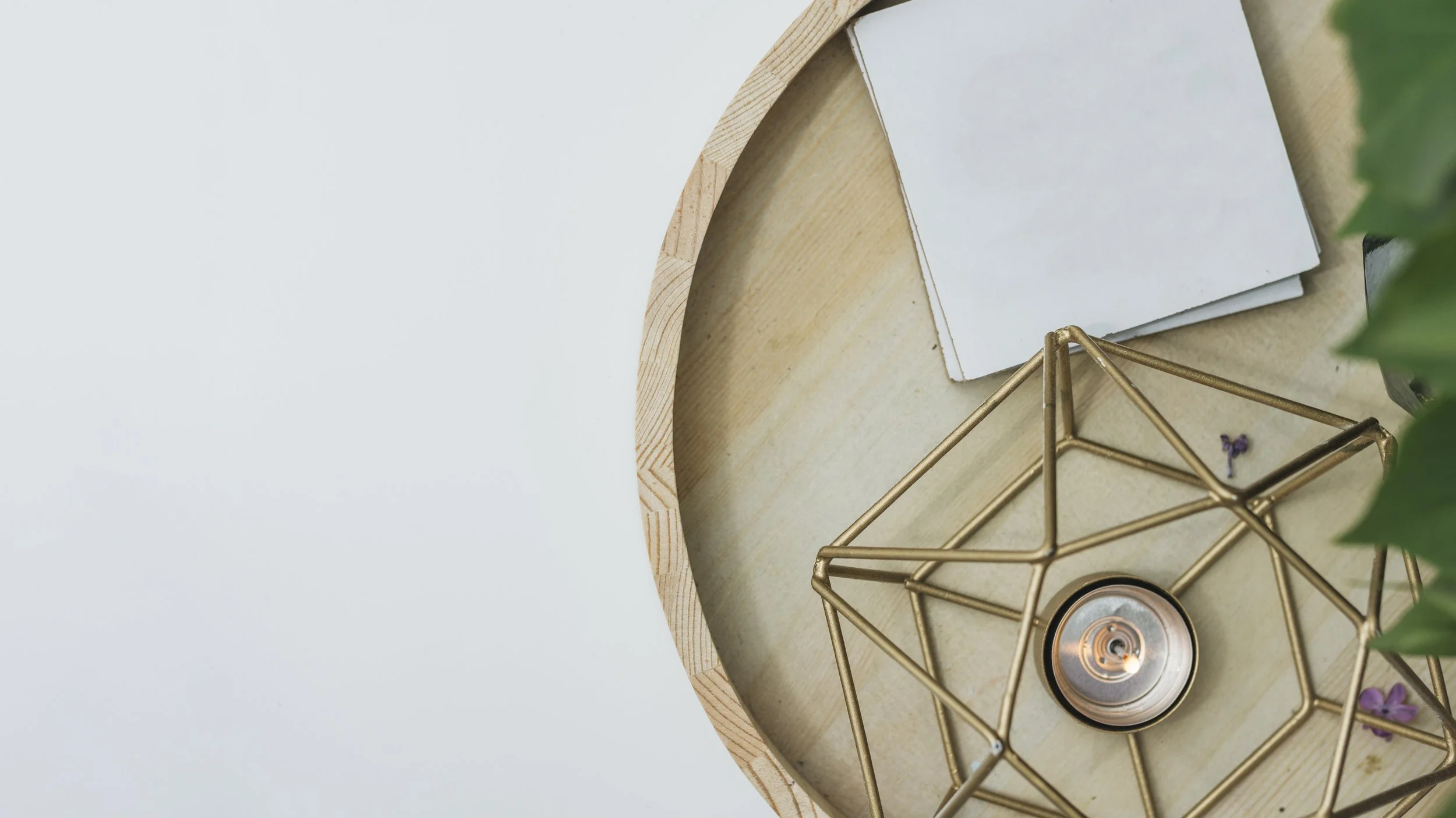 Top view of a round wooden tray with a white notepad, a brass geometric candle holder with a lit tealight, and some small purple flowers, with a green plant partially visible on the right side.