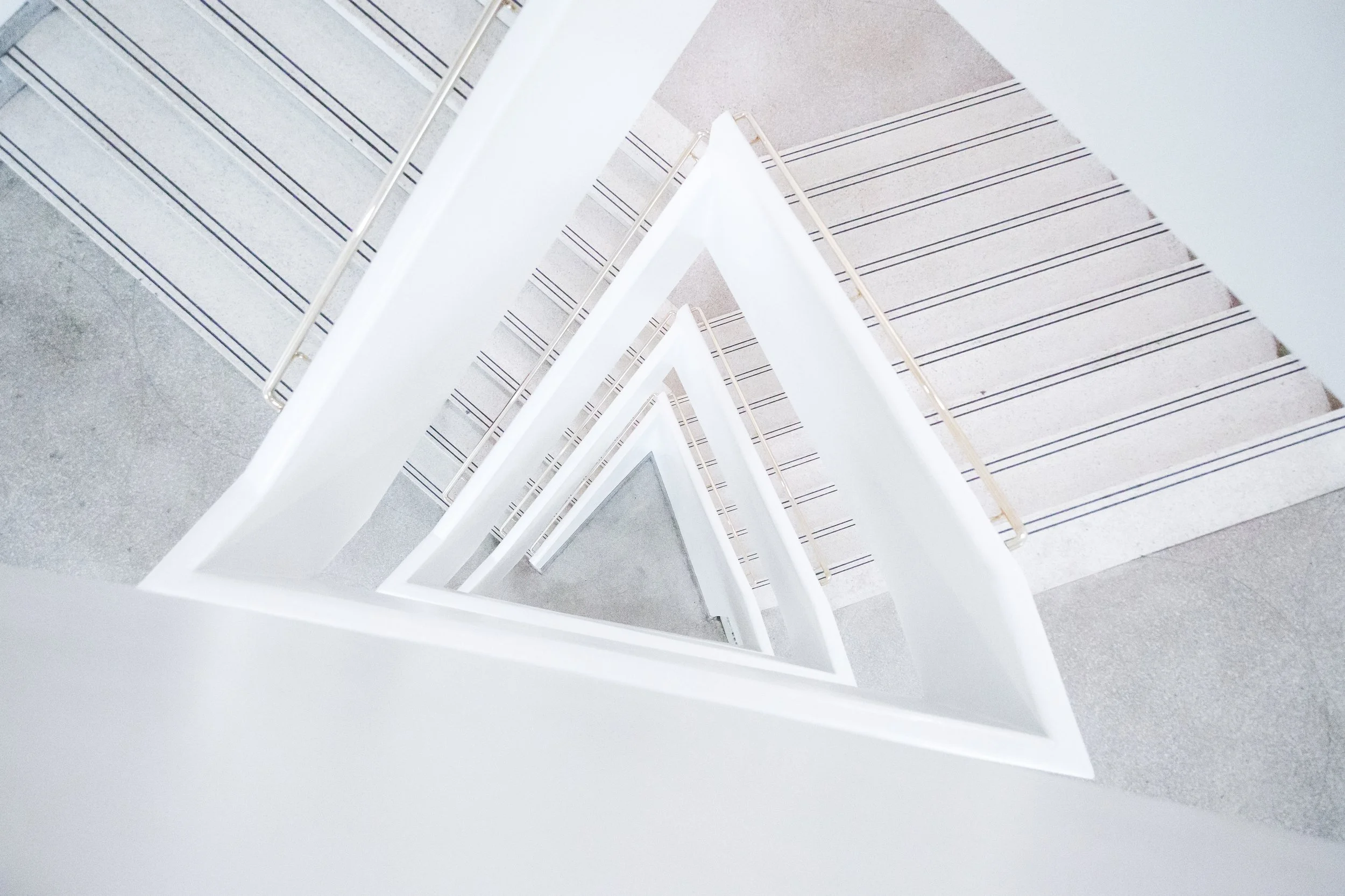 A white staircase viewed from above, with a triangular shape and metal handrails on both sides, descending in a zig-zag pattern across a building interior.