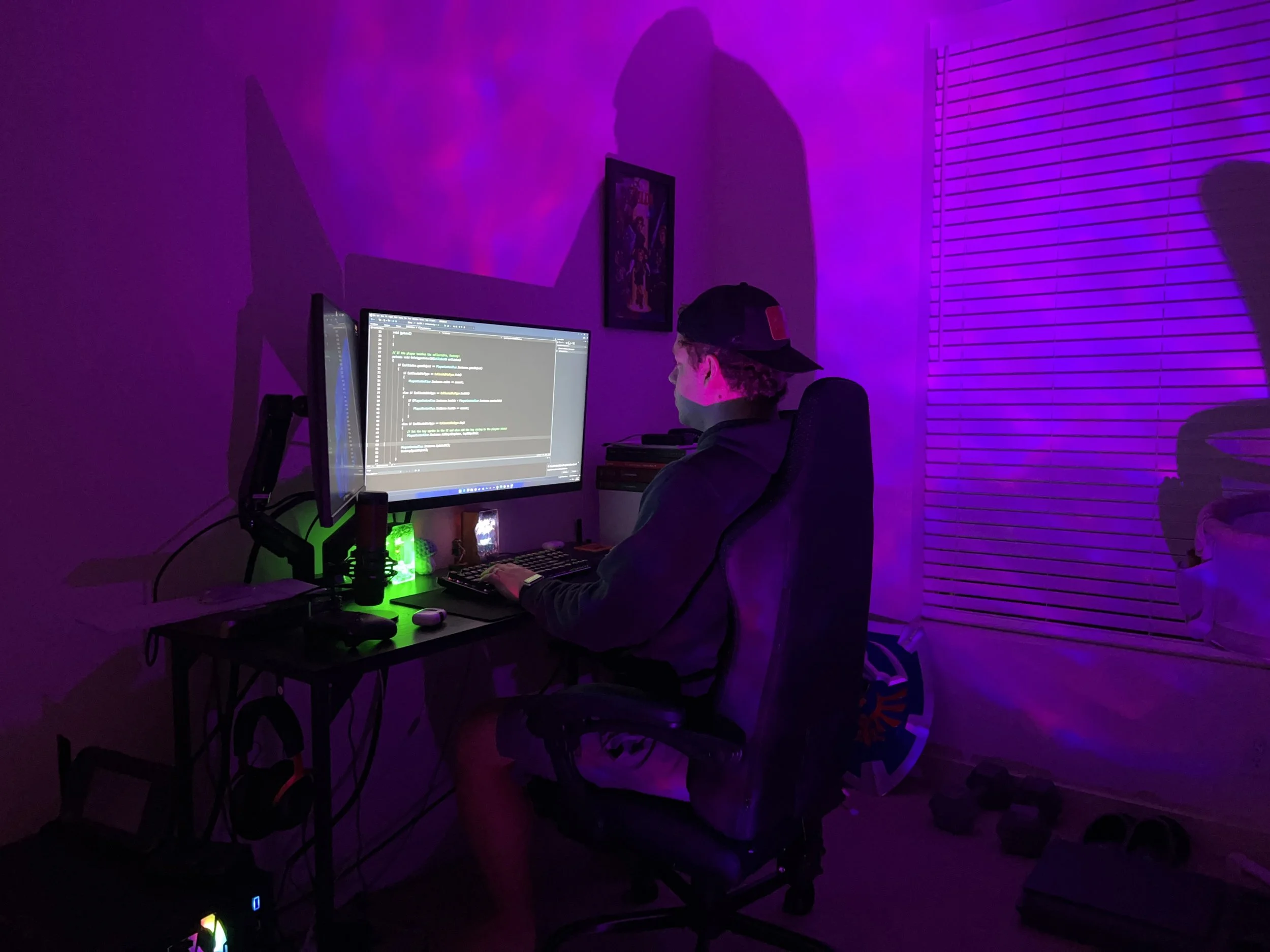 A person wearing a cap sitting at a desk with computer screens in a dark room illuminated by purple and green lights.
