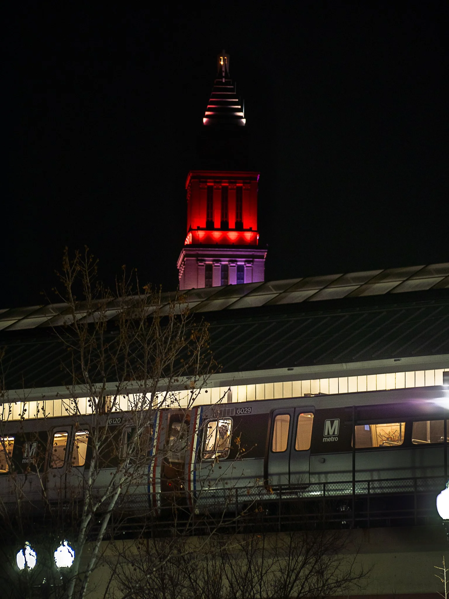 Night photo of a tall tower with red, purple, and white lighting, with a metro train on an elevated track in the foreground, and some bare trees and street lamps.