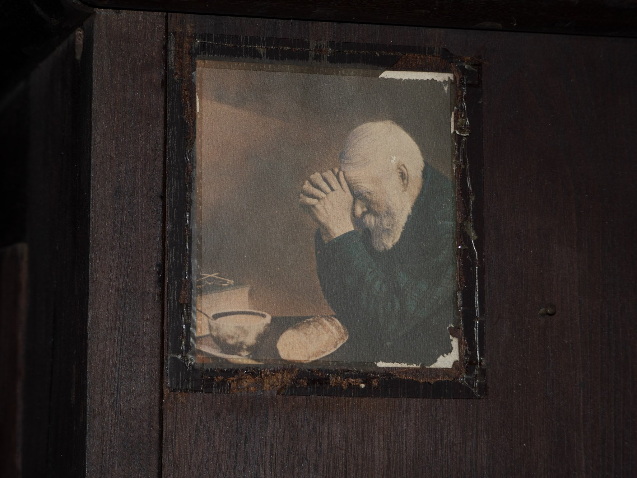 An old aging photograph of an elderly man with white hair and a beard, wearing a dark sweater, sitting with his hands clasped together near his face, and a bowl and bread in front of him.