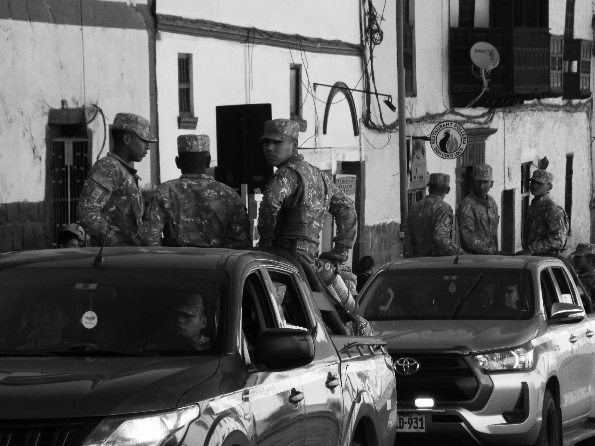 Group of soldiers in camouflage uniforms standing on a city street near parked cars.