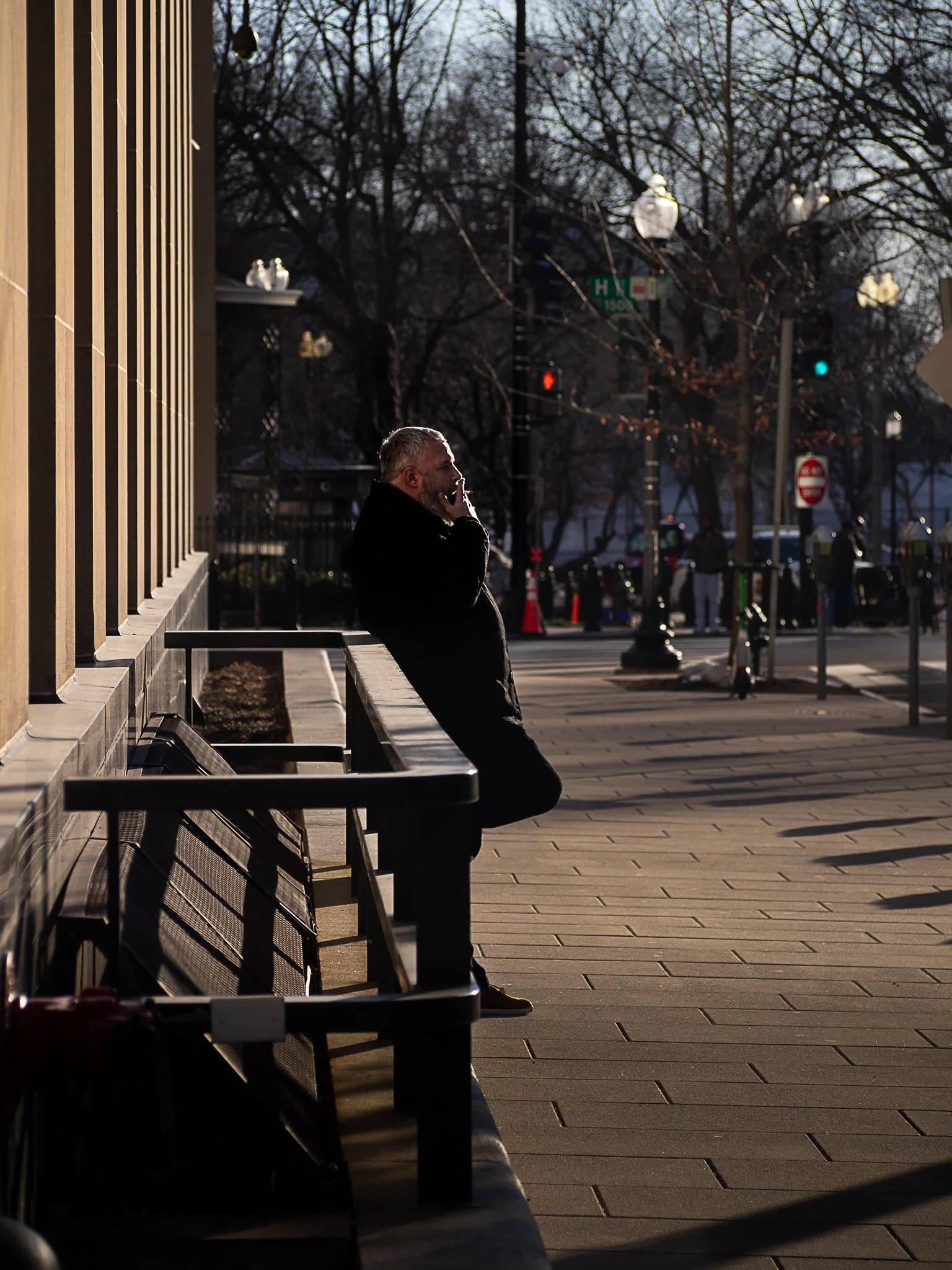 A man standing on a city sidewalk, leaning against a bench and talking on his cell phone, with trees and street signs in the background during sunset.