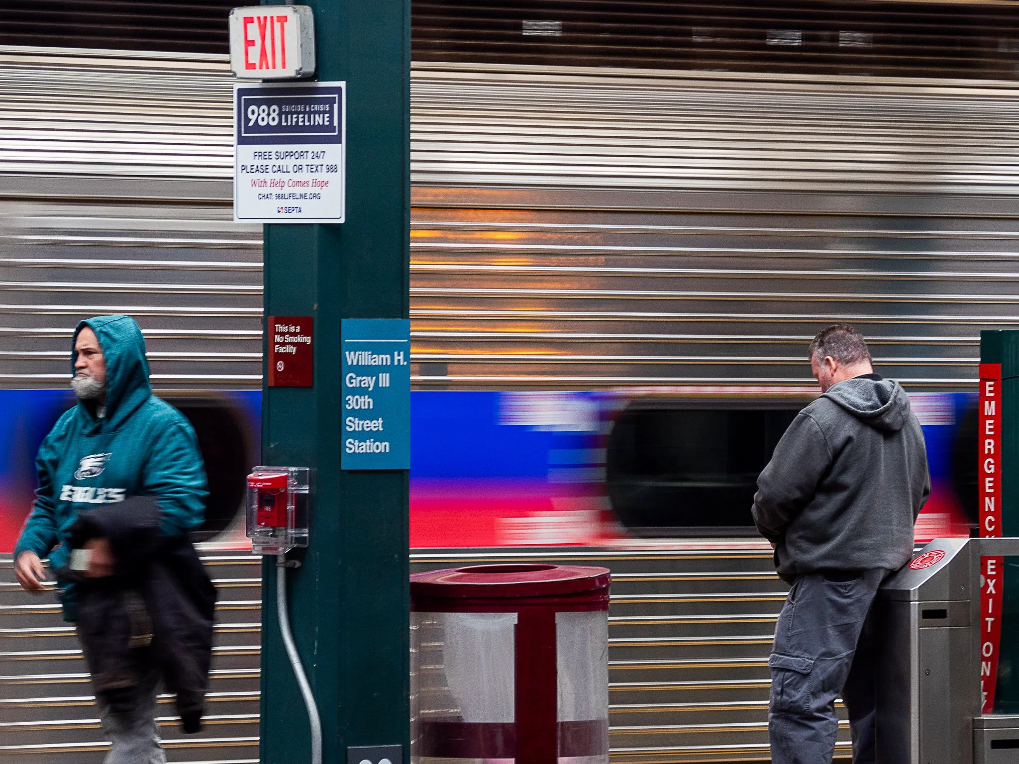 Two men at a subway station, one walking past wearing a teal Philadelphia Eagles hoodie and black pants, the other standing near the exit barrier wearing a gray hoodie and pants, with a moving train in the background.