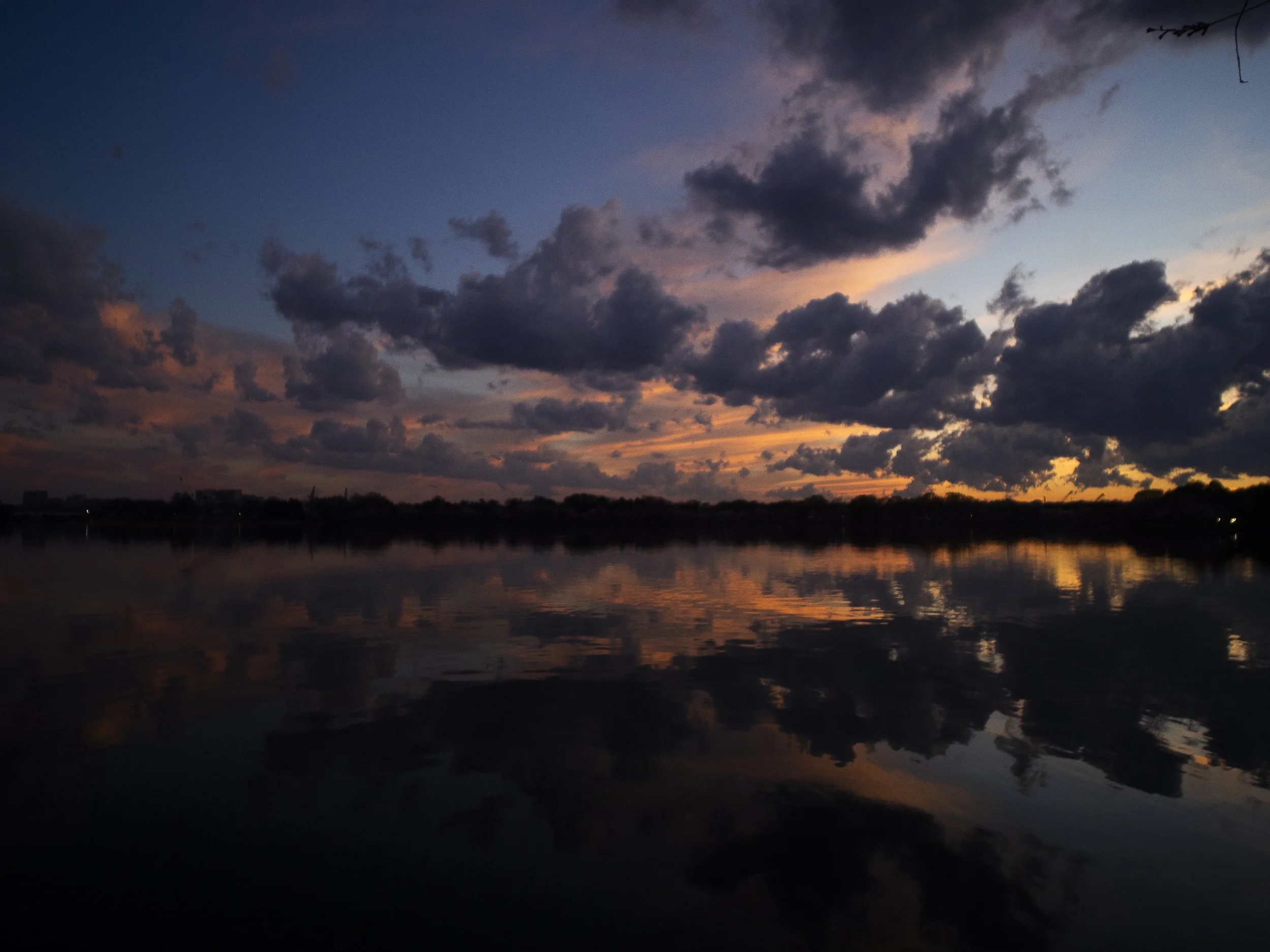 Sunset over a calm body of water with dark clouds in the sky and their reflections visible on the water surface.