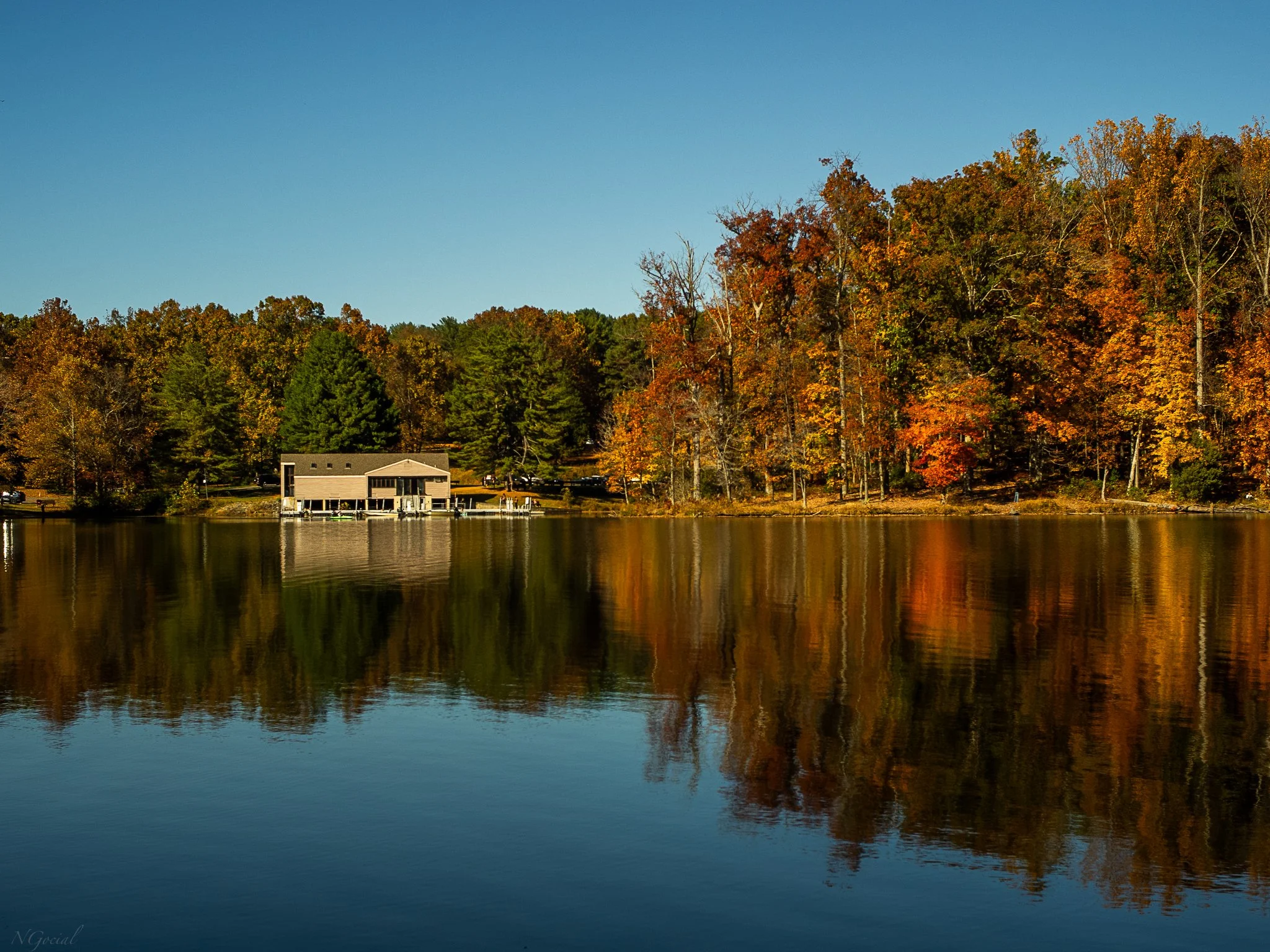 A lakeside scene with a boathouse and autumn trees reflected in calm water, under a clear blue sky.