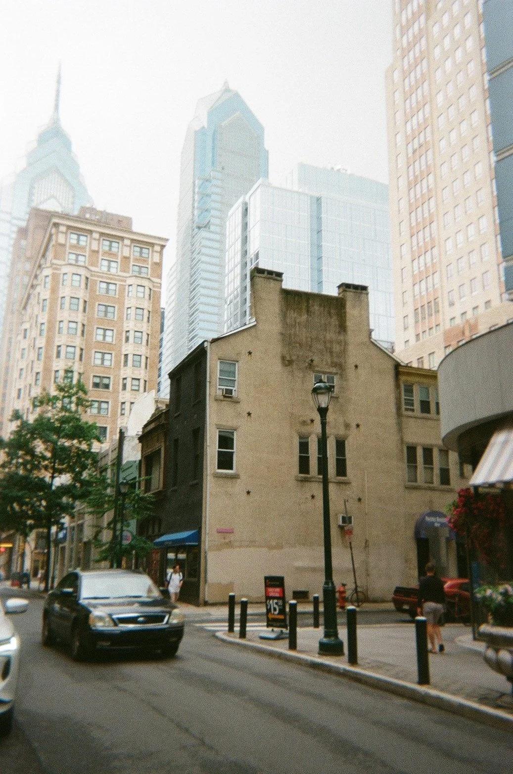 Urban street scene with historic beige building in foreground, surrounded by modern skyscrapers, trees, parked cars, and pedestrians.
