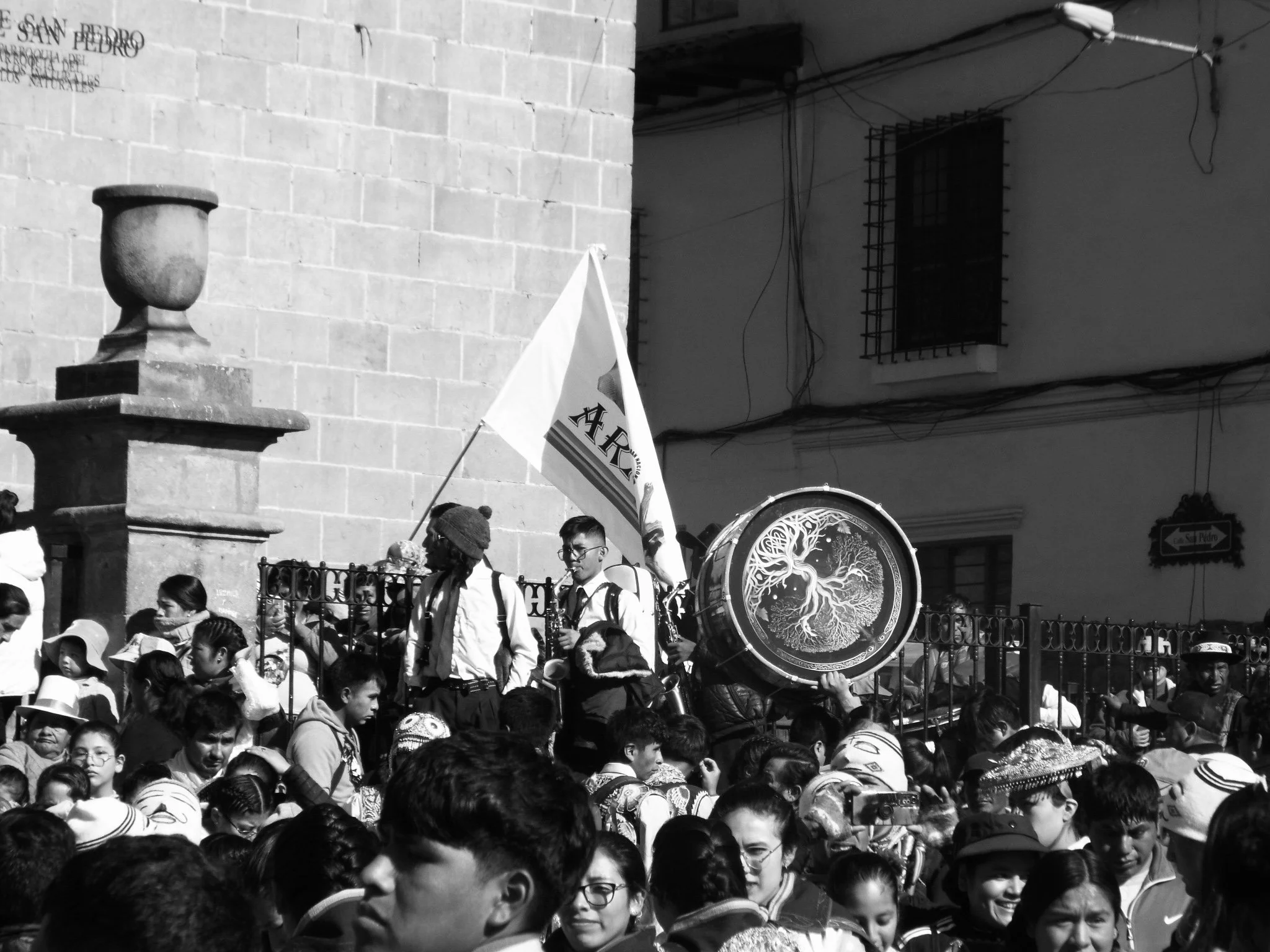 A crowd of people gathered at a street festival with a group of musicians playing instruments, a flag, and a large decorative drum.