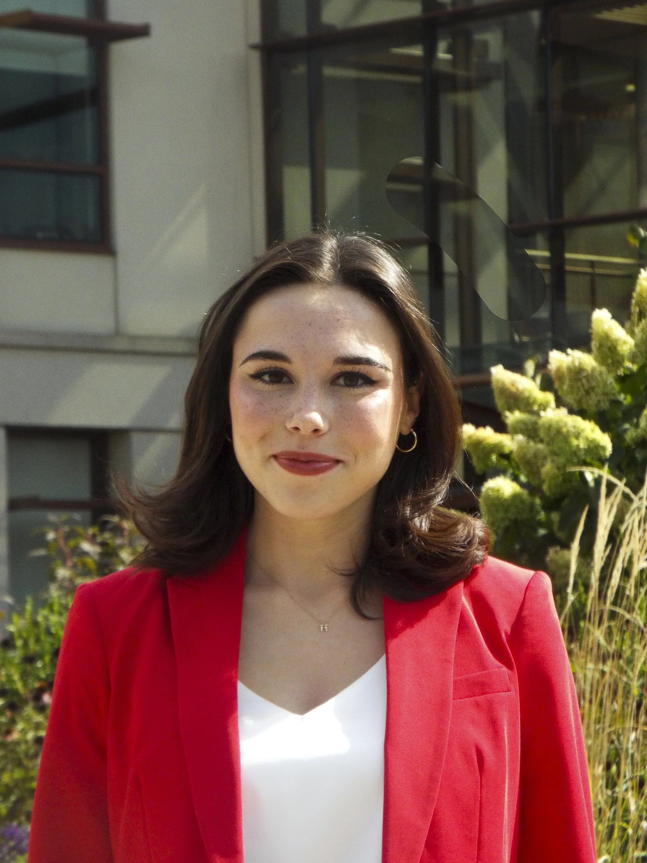 A woman with shoulder-length brown hair, wearing a red blazer and white top, standing outdoors near greenery and a modern glass building.