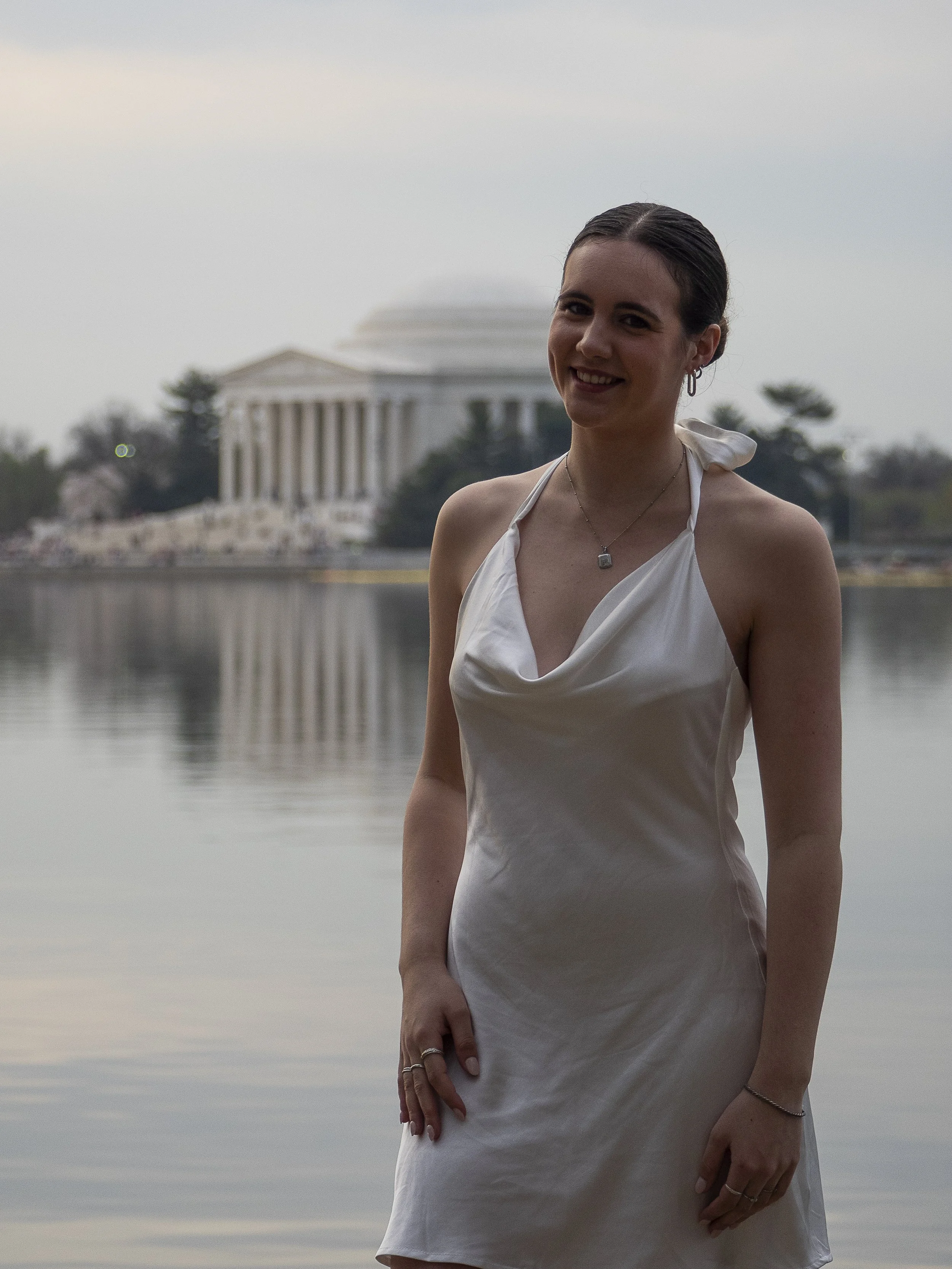 A young woman in a white dress standing outdoors near a body of water with the Lincoln Memorial in Washington, D.C., in the background.