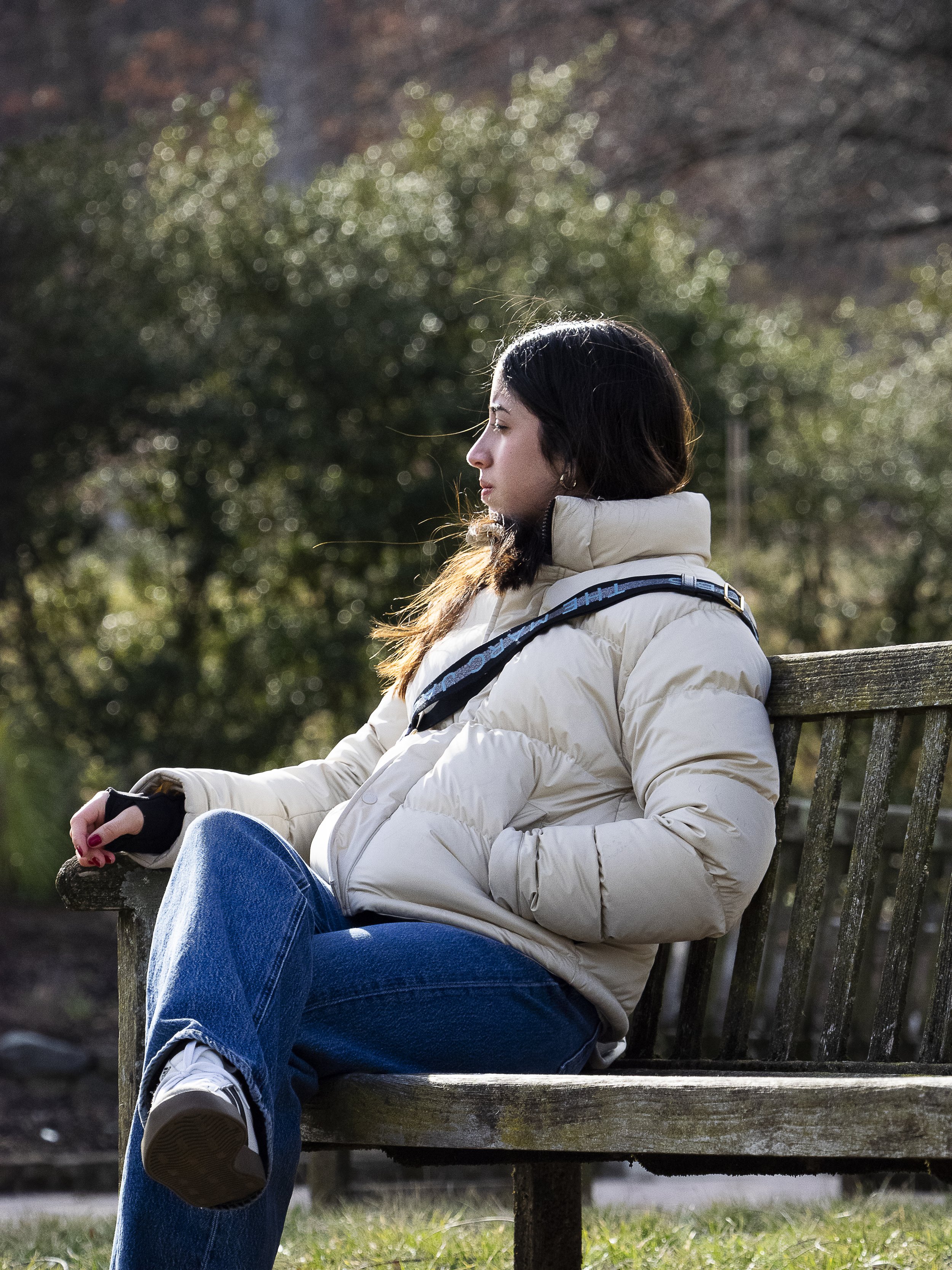 A woman sitting on a wooden park bench, wearing a beige puffer jacket, blue jeans, and white sneakers, with her eyes closed and head slightly tilted down, surrounded by greenery and trees.