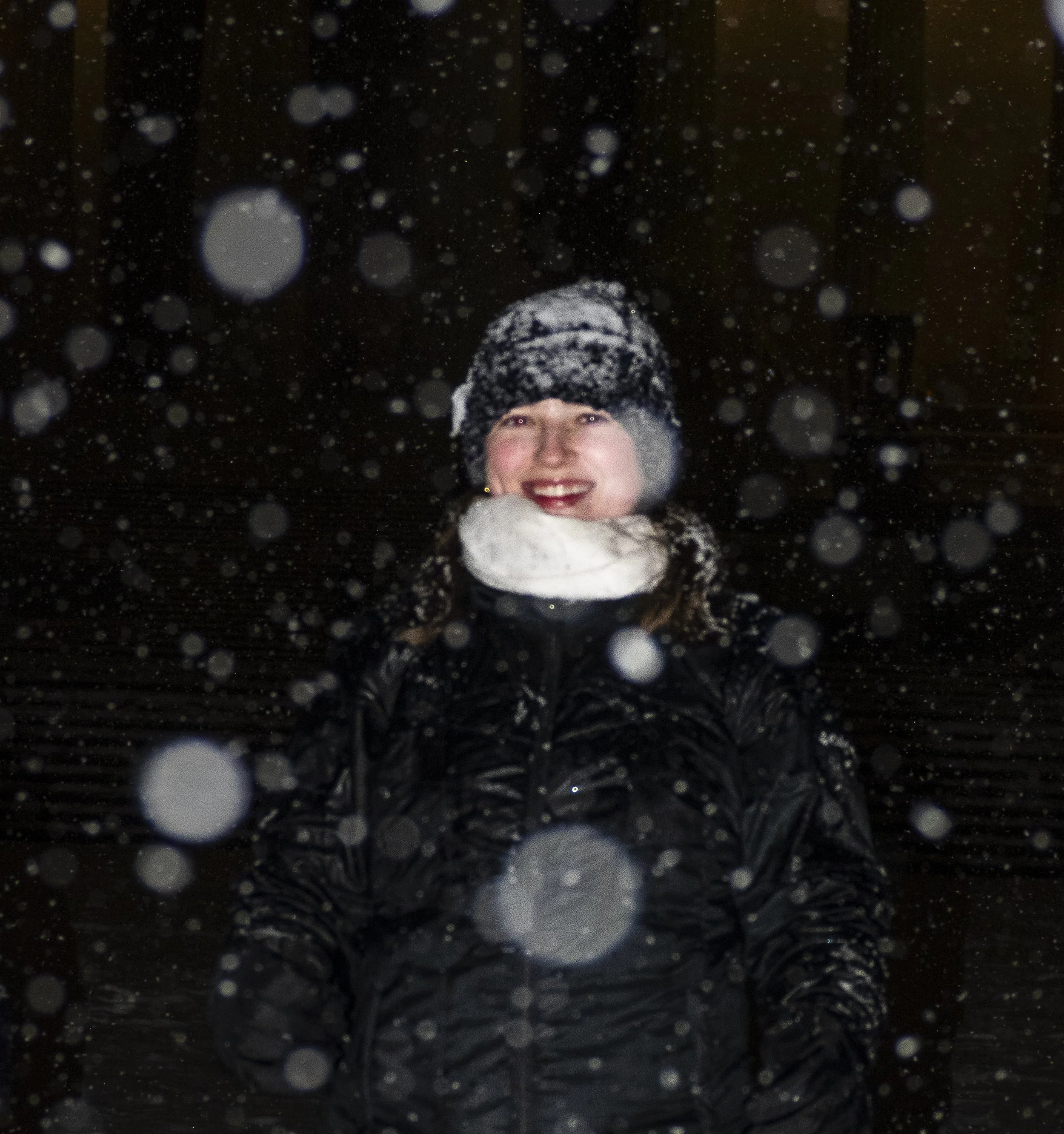 A woman smiling outdoors at night in heavy snowfall, wearing a black winter jacket, a black and gray knitted hat, and a white scarf.