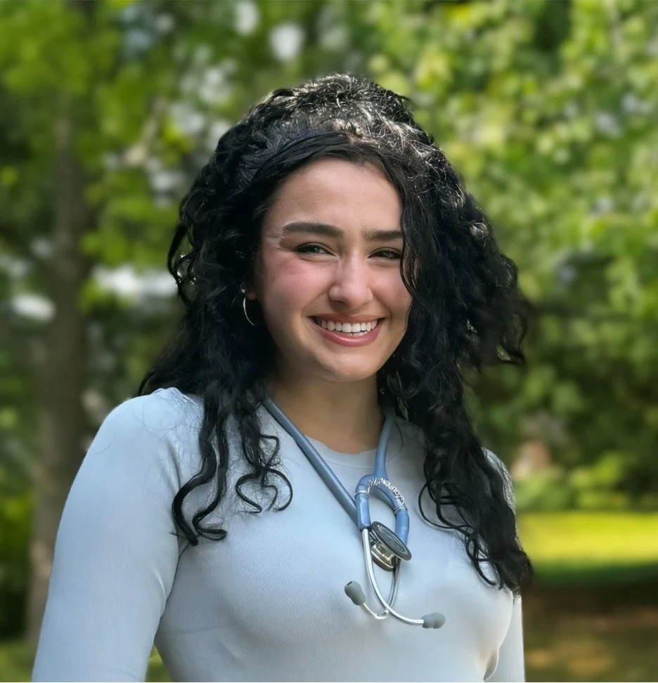 A smiling person with long curly hair wearing a light blue top and a stethoscope around their neck, standing outdoors with greenery in the background.